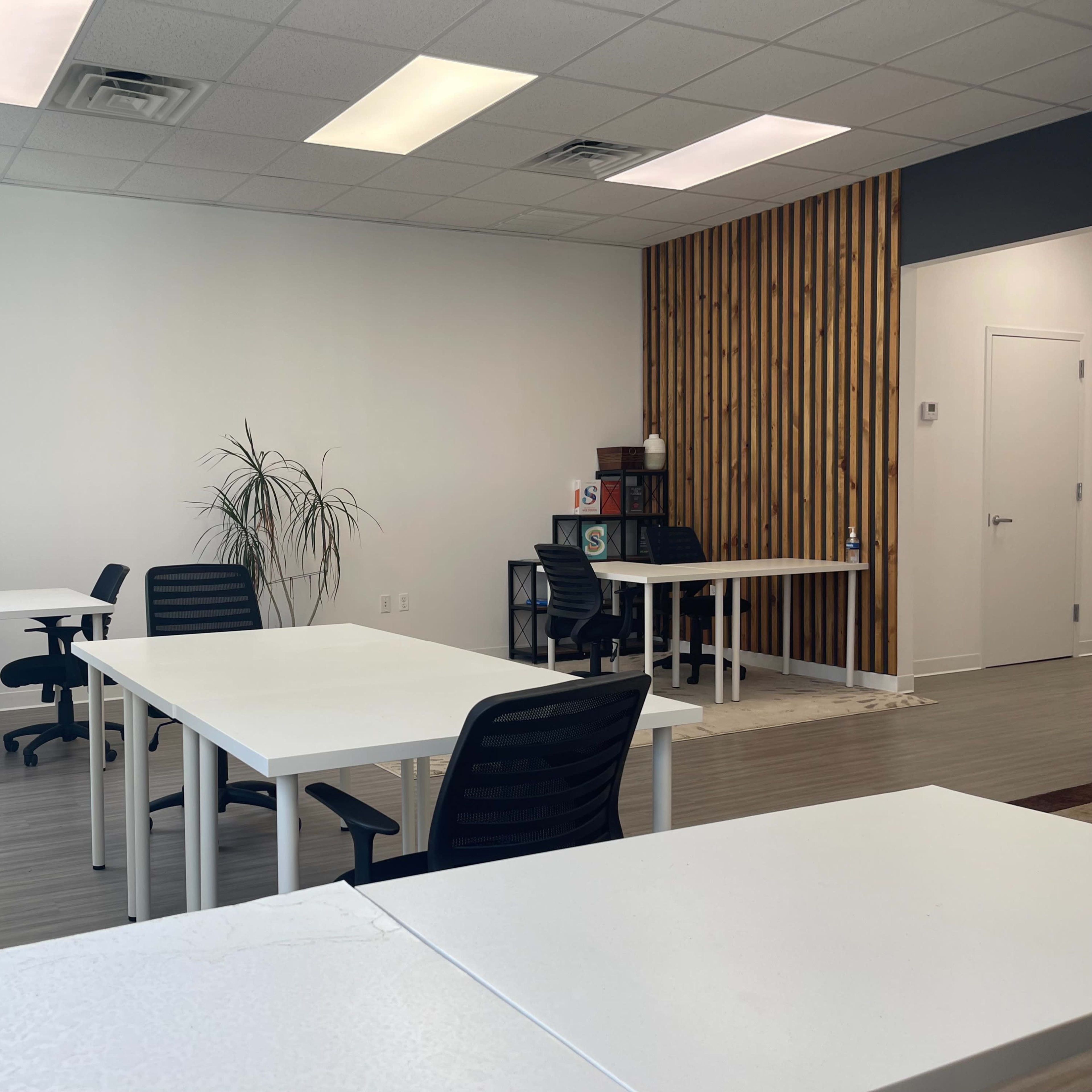 A spacious office with white tables and black chairs, a large window, and a wooden accent wall.