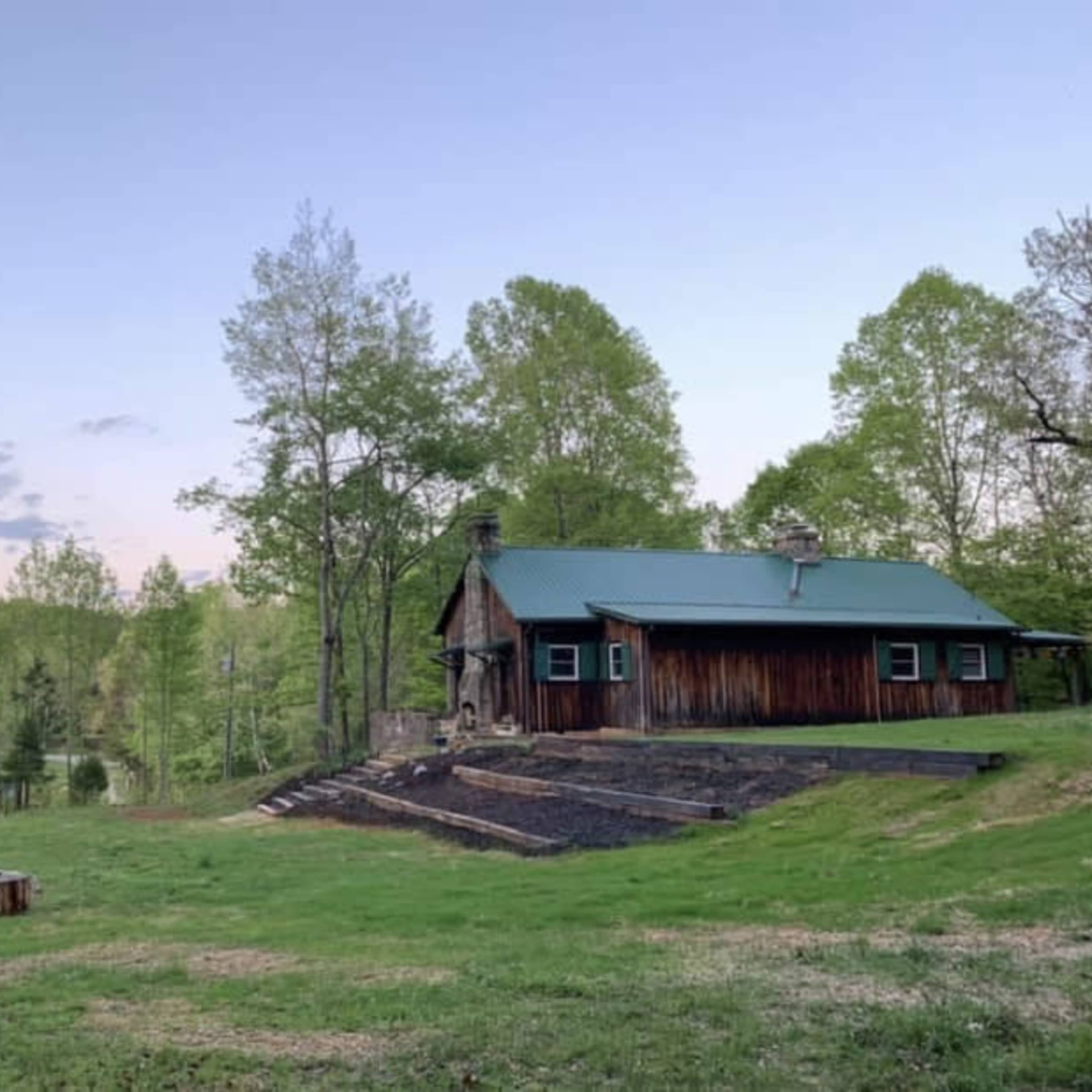 A wooden cabin with a green metal roof sits on a grassy hillside surrounded by trees and a fire pit area.