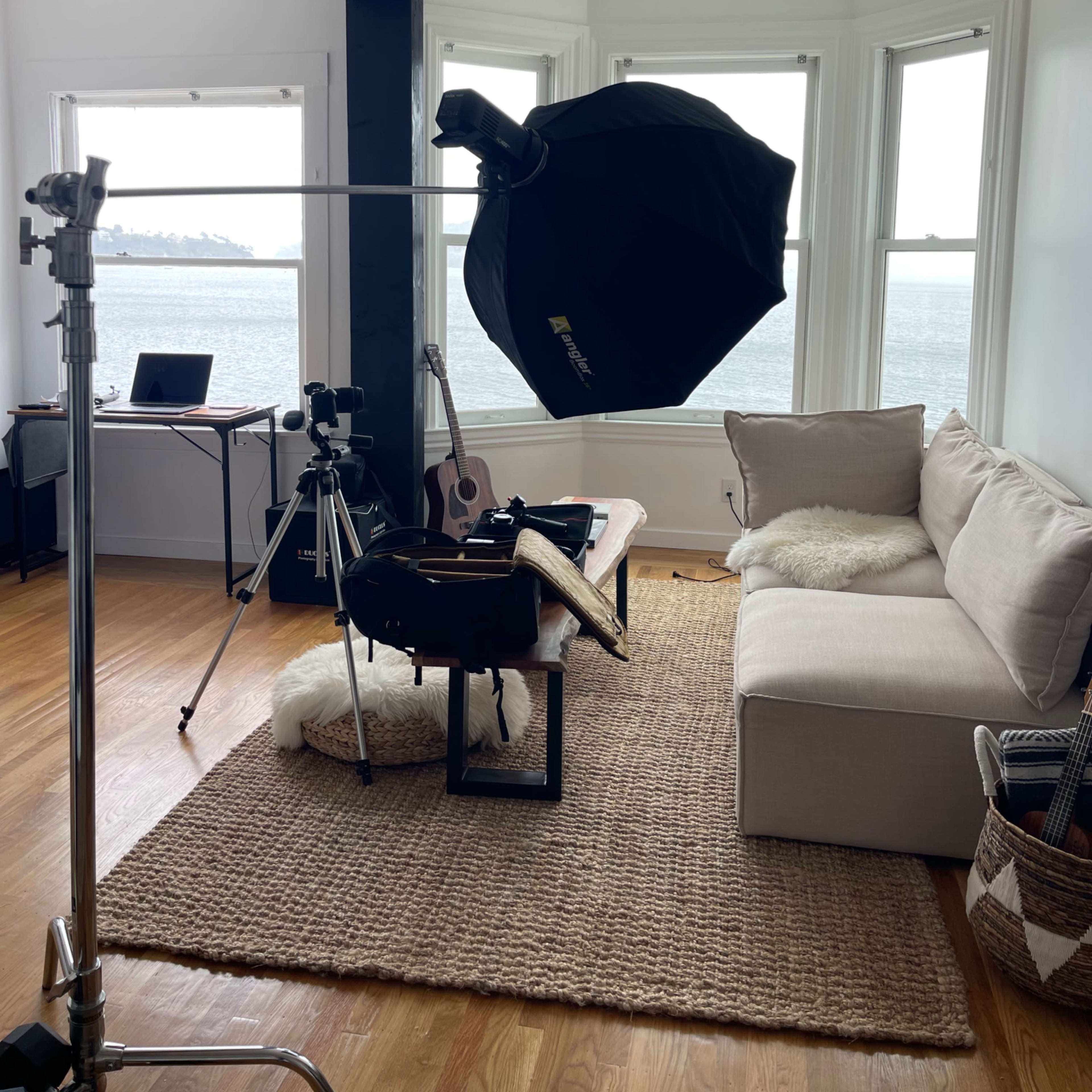 A photography setup is arranged in a sunny room with panoramic windows, featuring a softbox lighting equipment, a couch, and various instruments and equipment on a table.