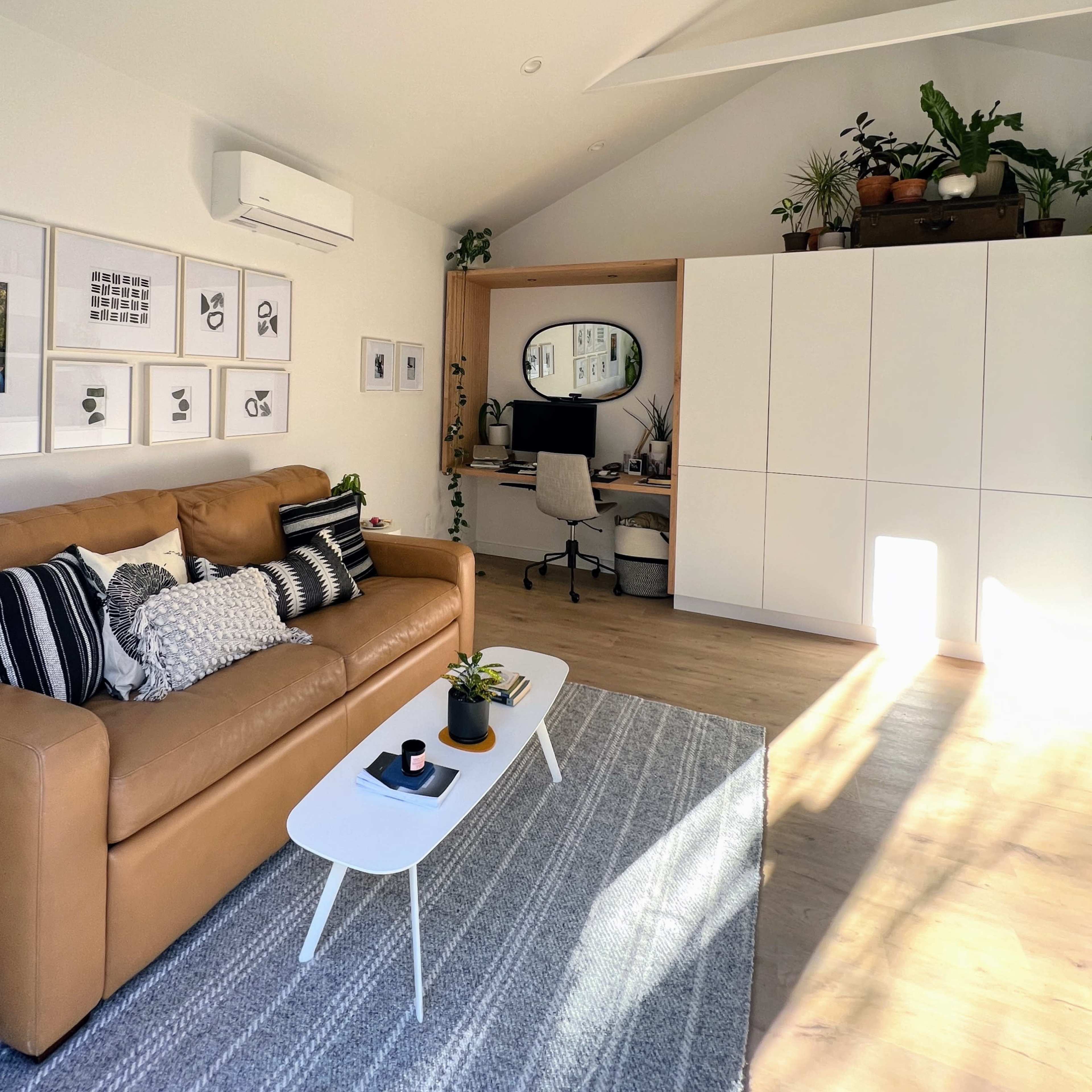A modern living area with a tan couch, a white coffee table, and a workspace featuring a desk and plants against a light-colored wall.