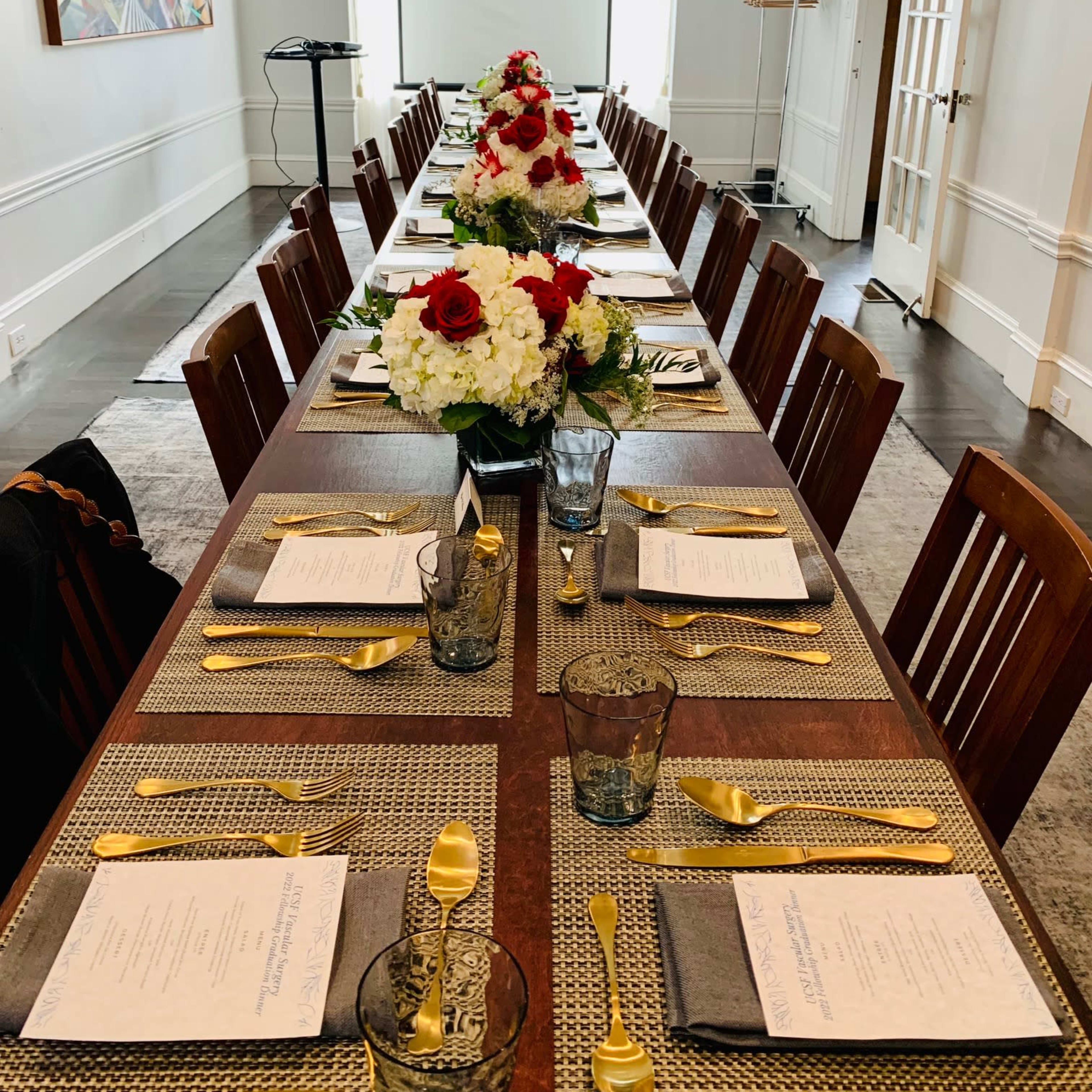 A long dining table is set with elegant place settings and floral centerpieces, arranged in a brightly lit room with a projector screen in the background.
