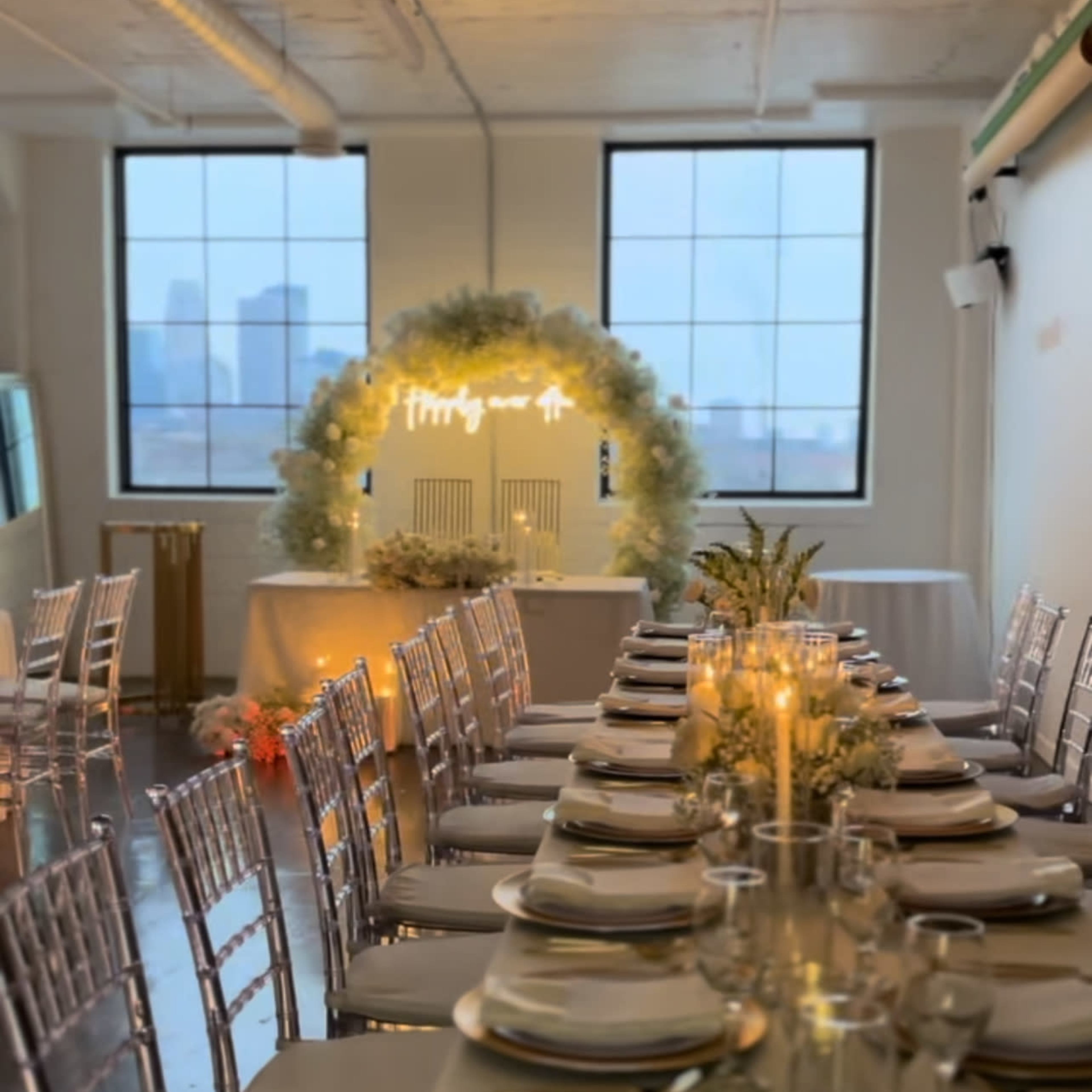 A long banquet table is set with elegant tableware in a well-lit room featuring large windows overlooking a city skyline.