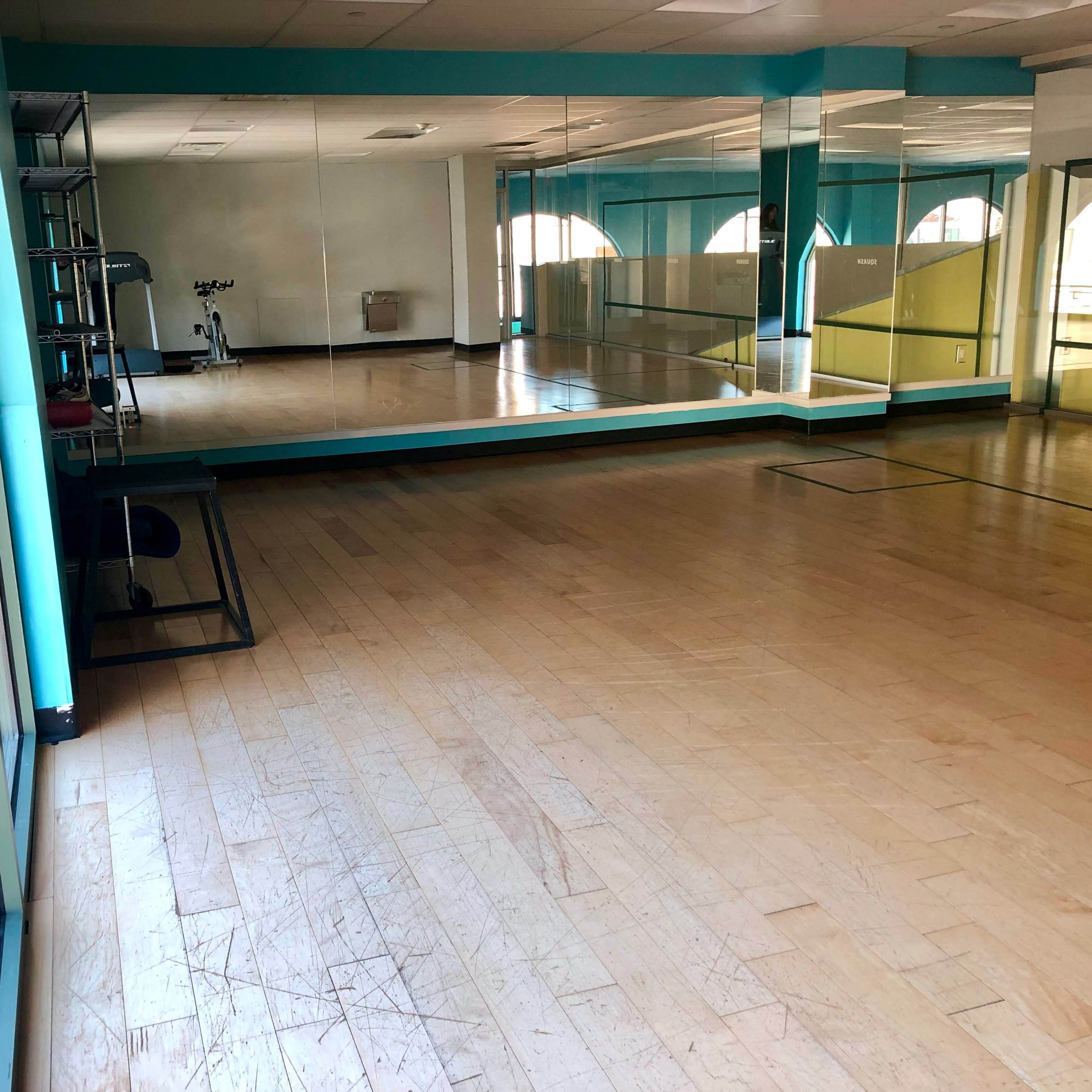 An empty fitness studio with wooden flooring and large mirrors on the walls, illuminated by natural light from windows.