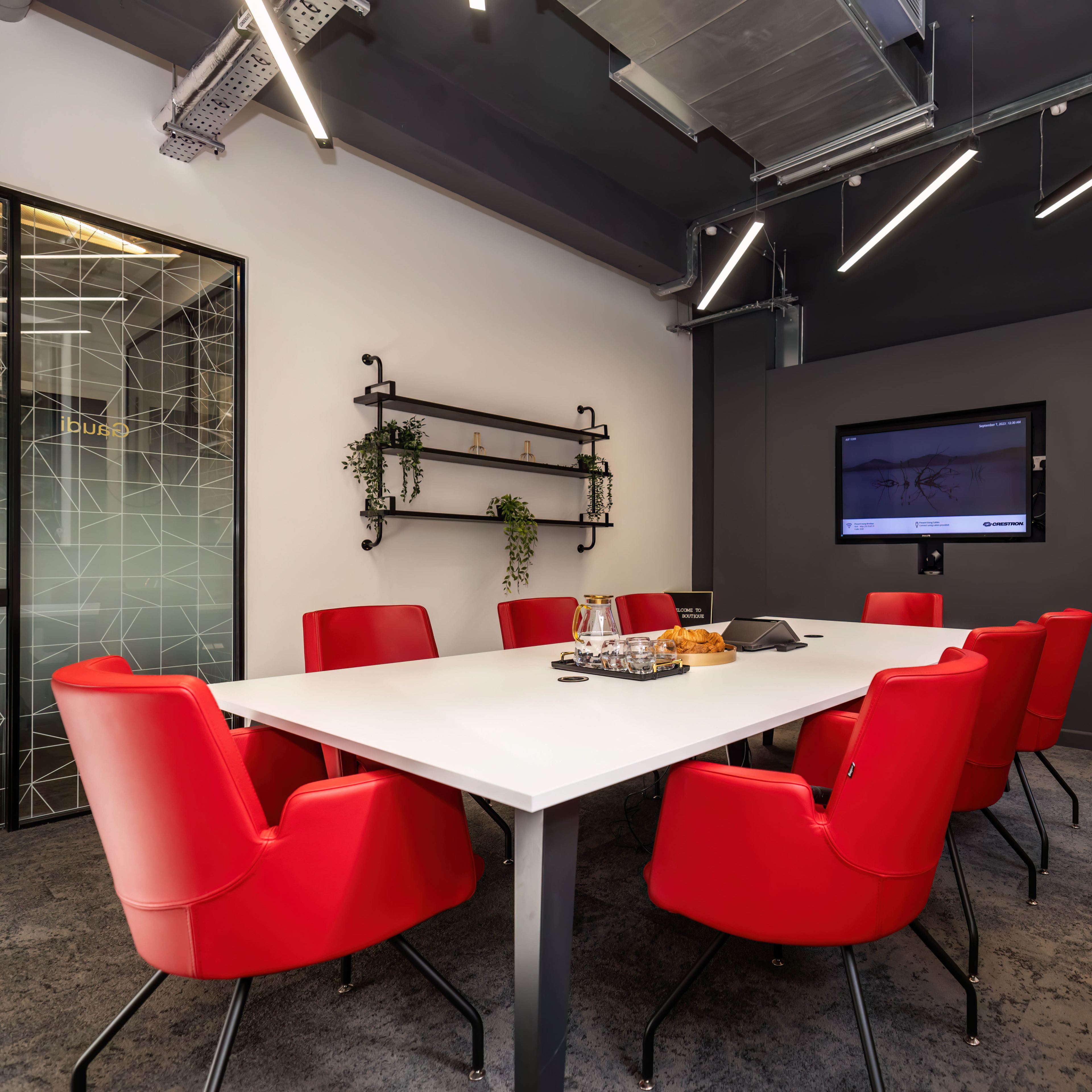 A conference room features a large white table surrounded by red chairs, with a large screen and shelves displaying plants on the walls.