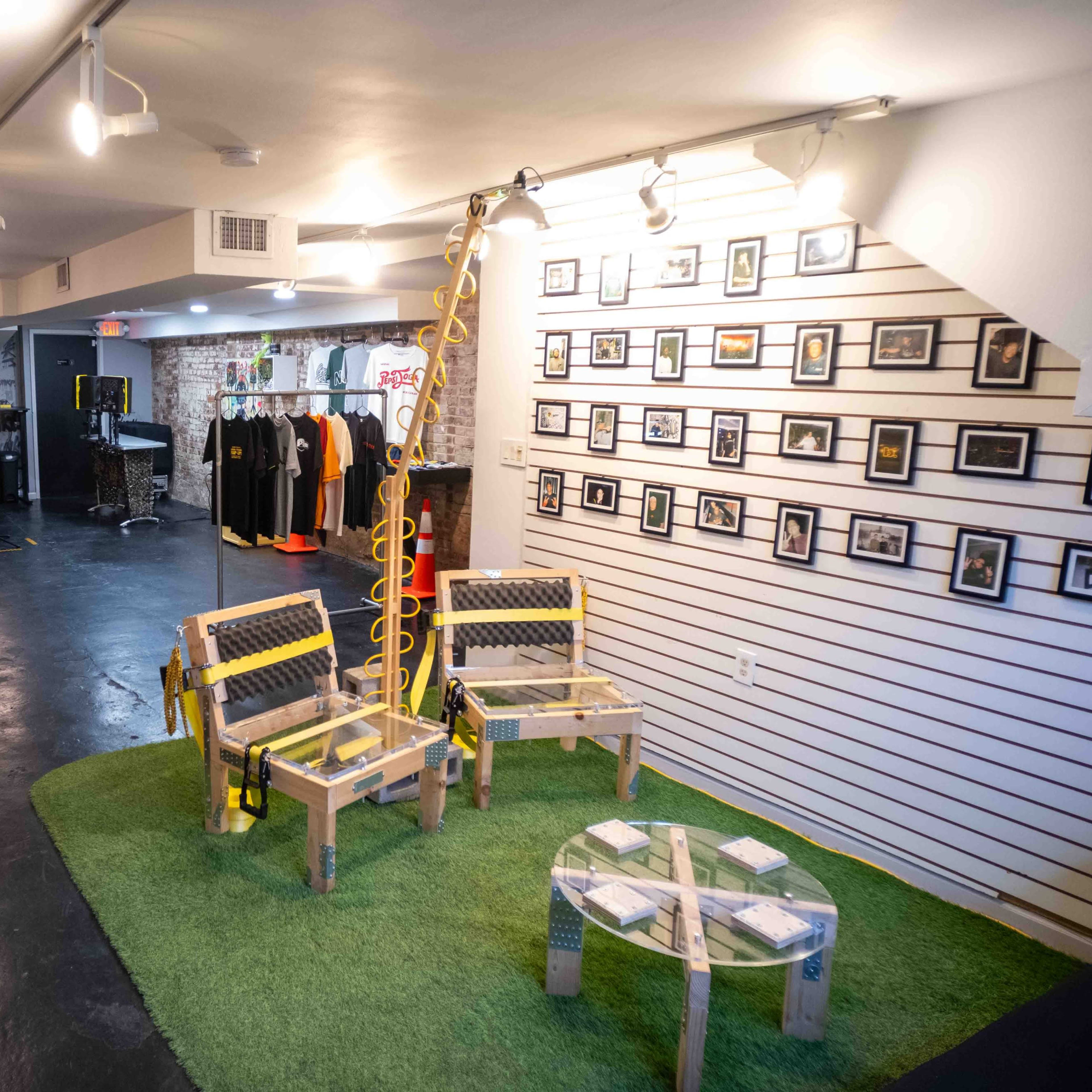 An indoor space with a brick wall, wooden chairs with yellow straps, a green carpet area, and photo frames displayed on a white panel, alongside clothing racks.