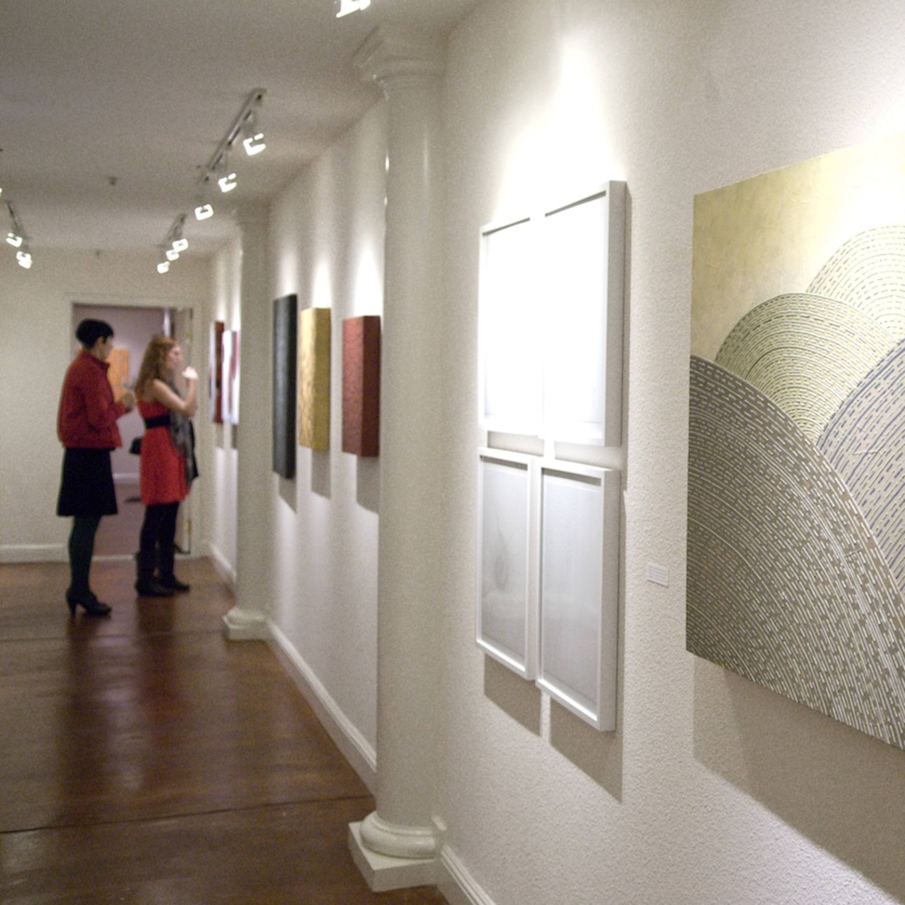 An art gallery corridor with a large abstract painting of hills on one wall and two people conversing in the background.