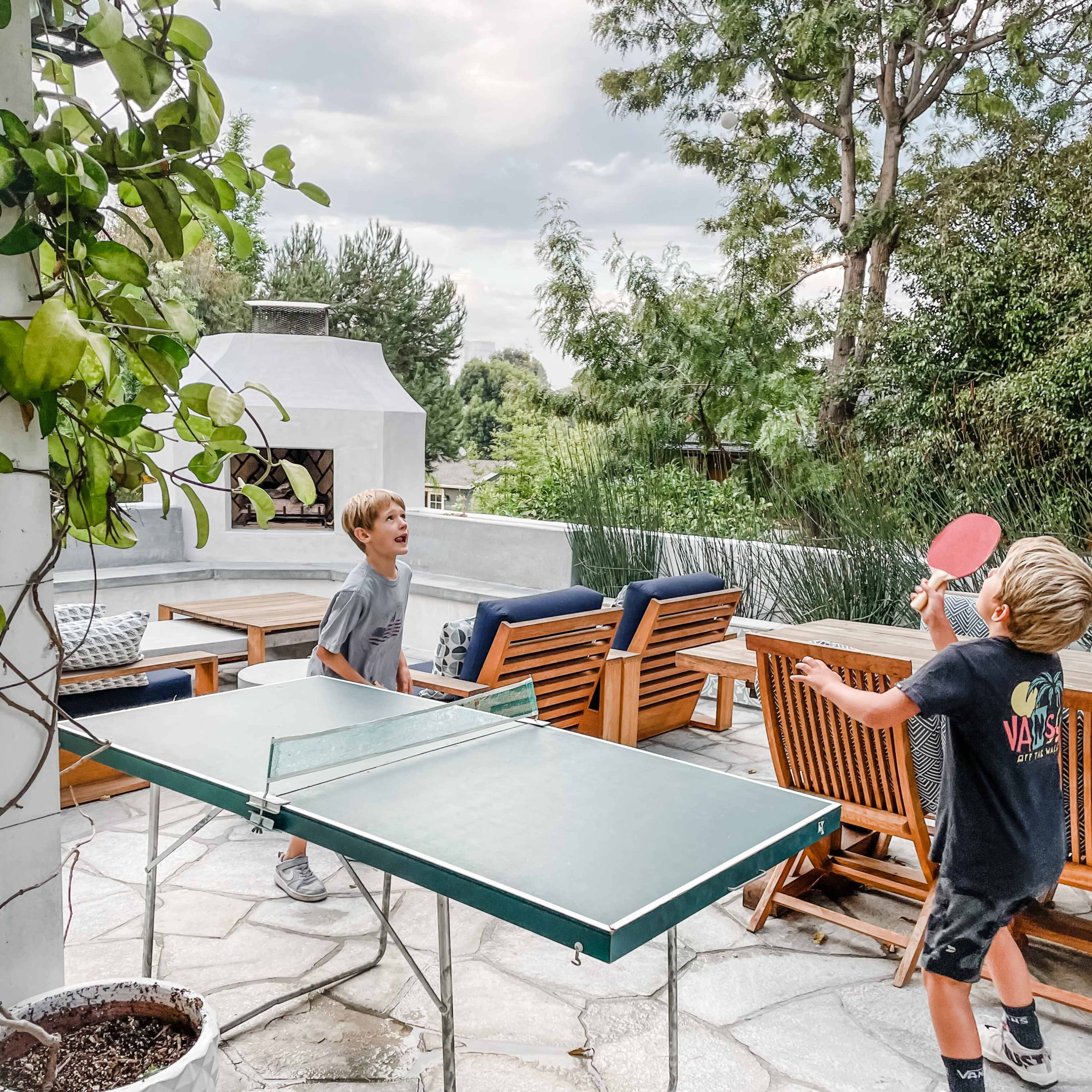 Two boys play table tennis on a patio surrounded by greenery and outdoor seating.