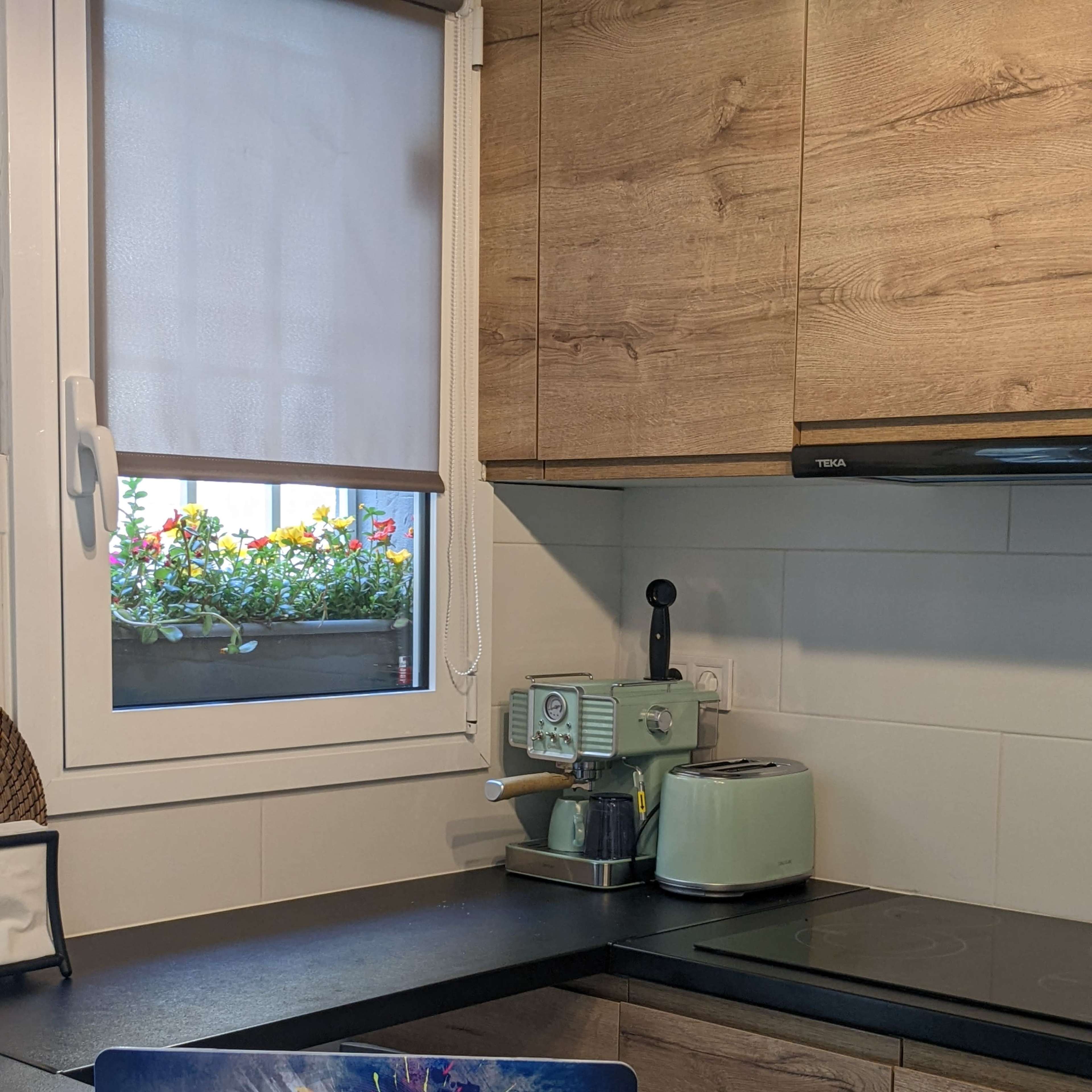 A laptop with a colorful design is placed on a black countertop in a kitchen, featuring a window with potted flowers and various kitchen appliances nearby.