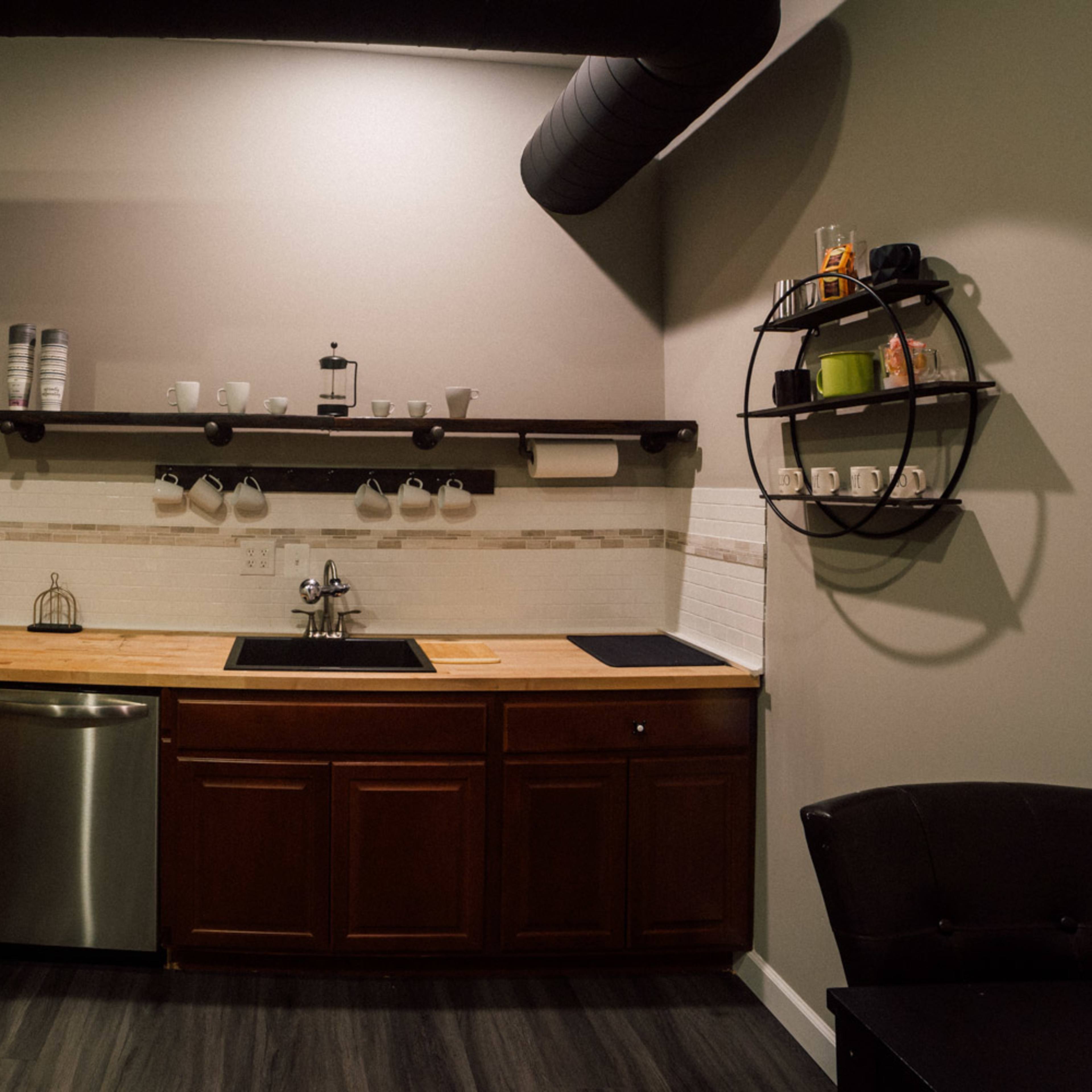 A modern kitchen area with a wood countertop, stainless steel sink, and shelving displaying mugs and a plant.