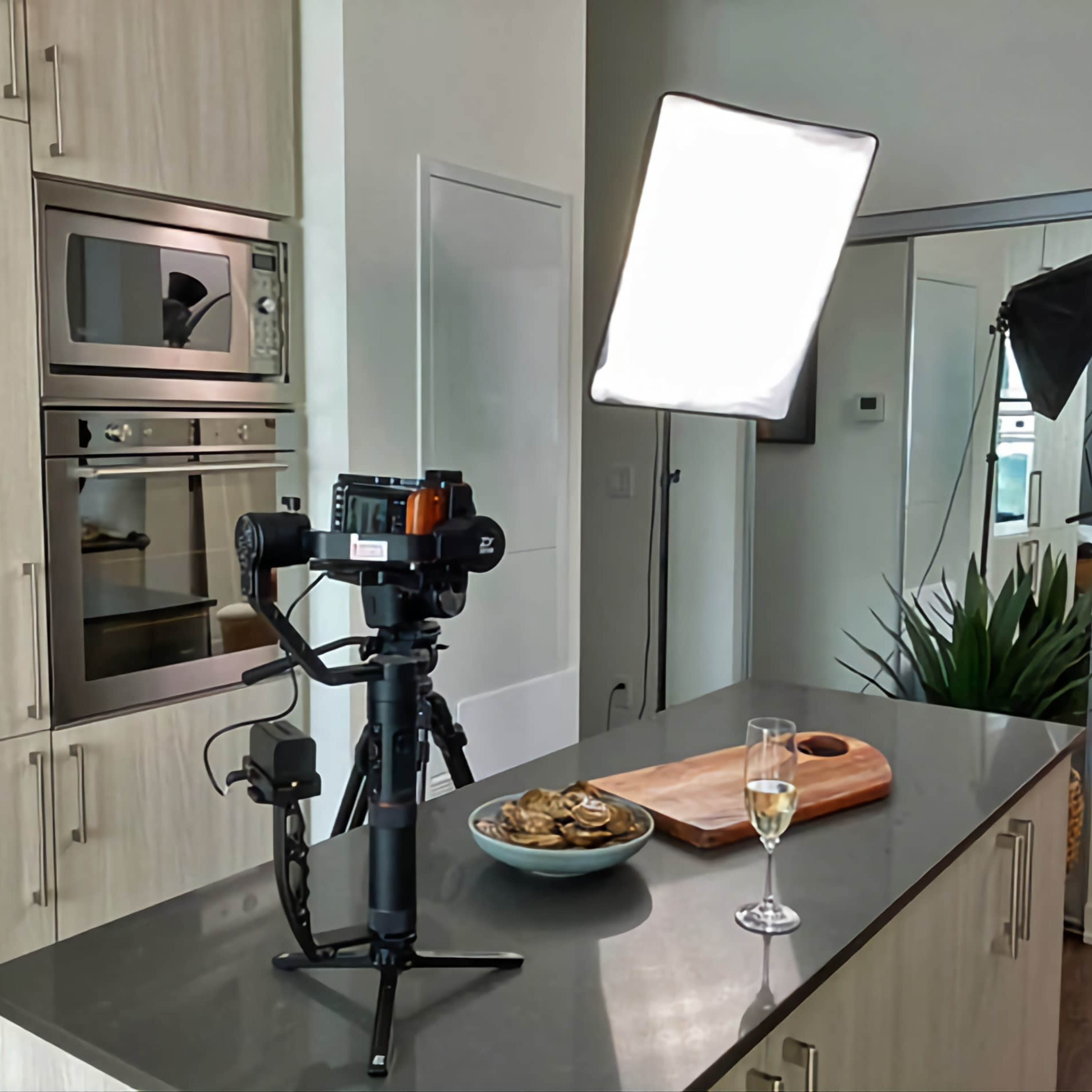 A camera on a tripod is set up on a kitchen countertop next to a glass of champagne and a bowl of food, with soft lighting equipment in the background.