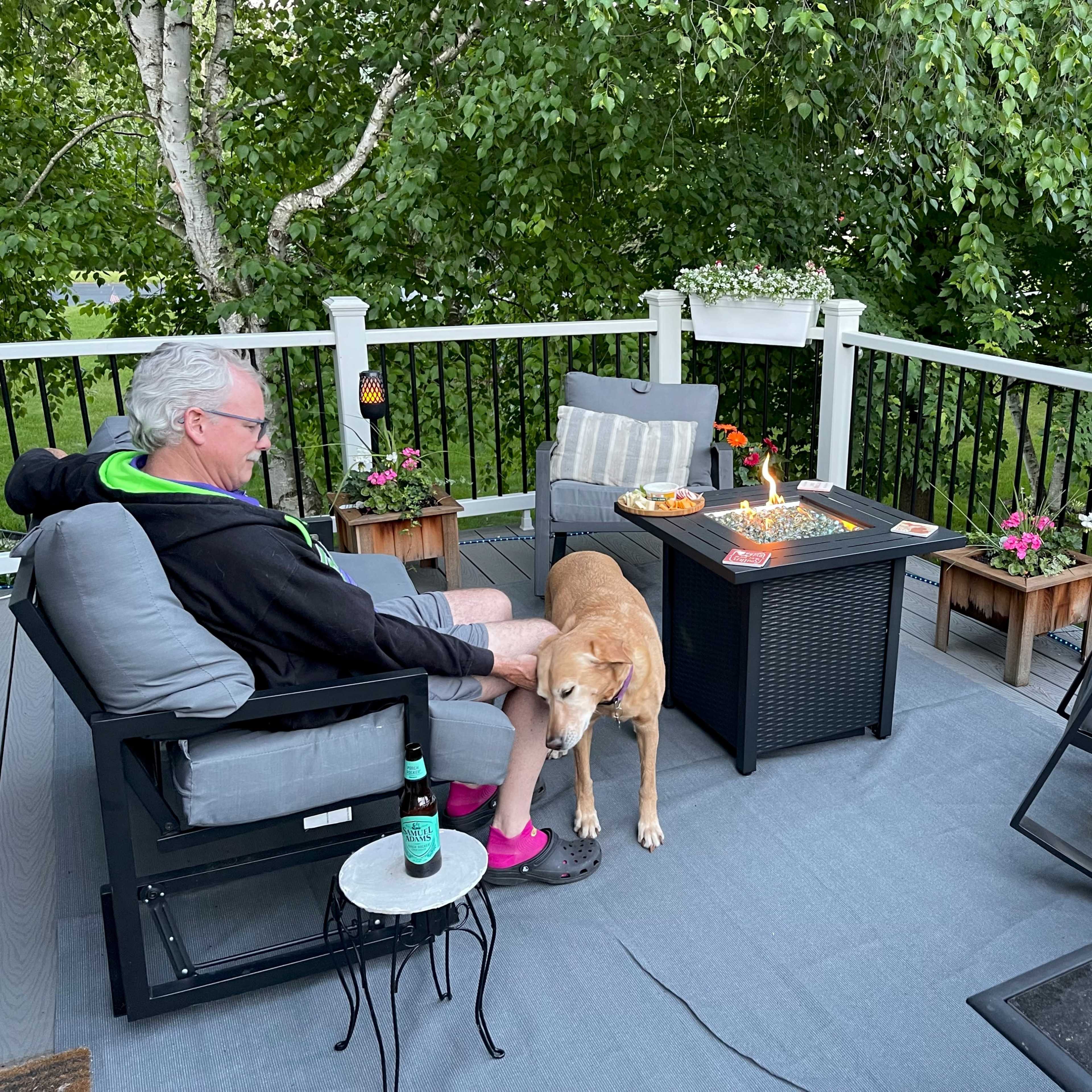 A man sits on a deck with a dog by his side, surrounded by potted plants and snacks on a table.