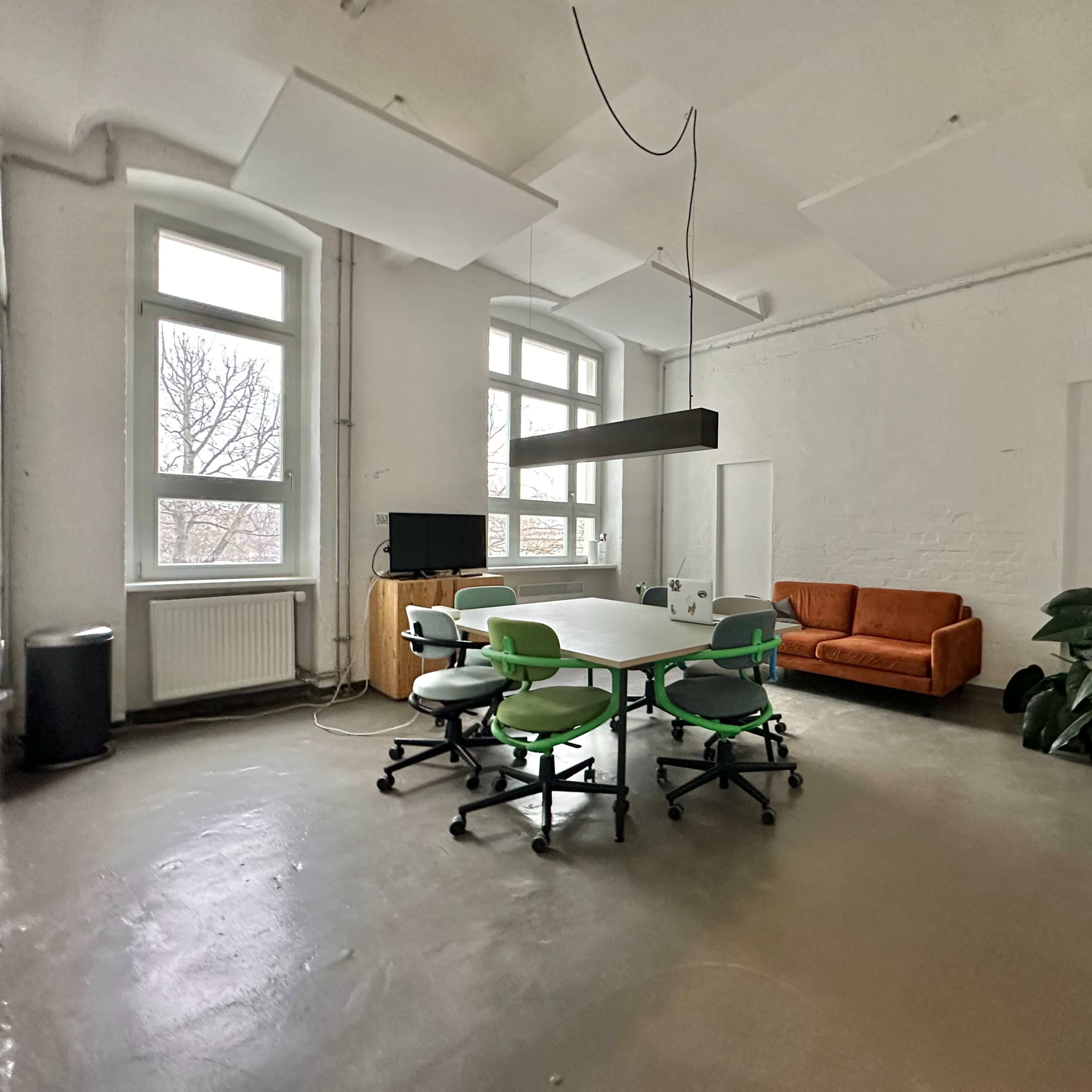 A spacious room features a central meeting table surrounded by green office chairs, an orange sofa, a television, and large windows allowing natural light.
