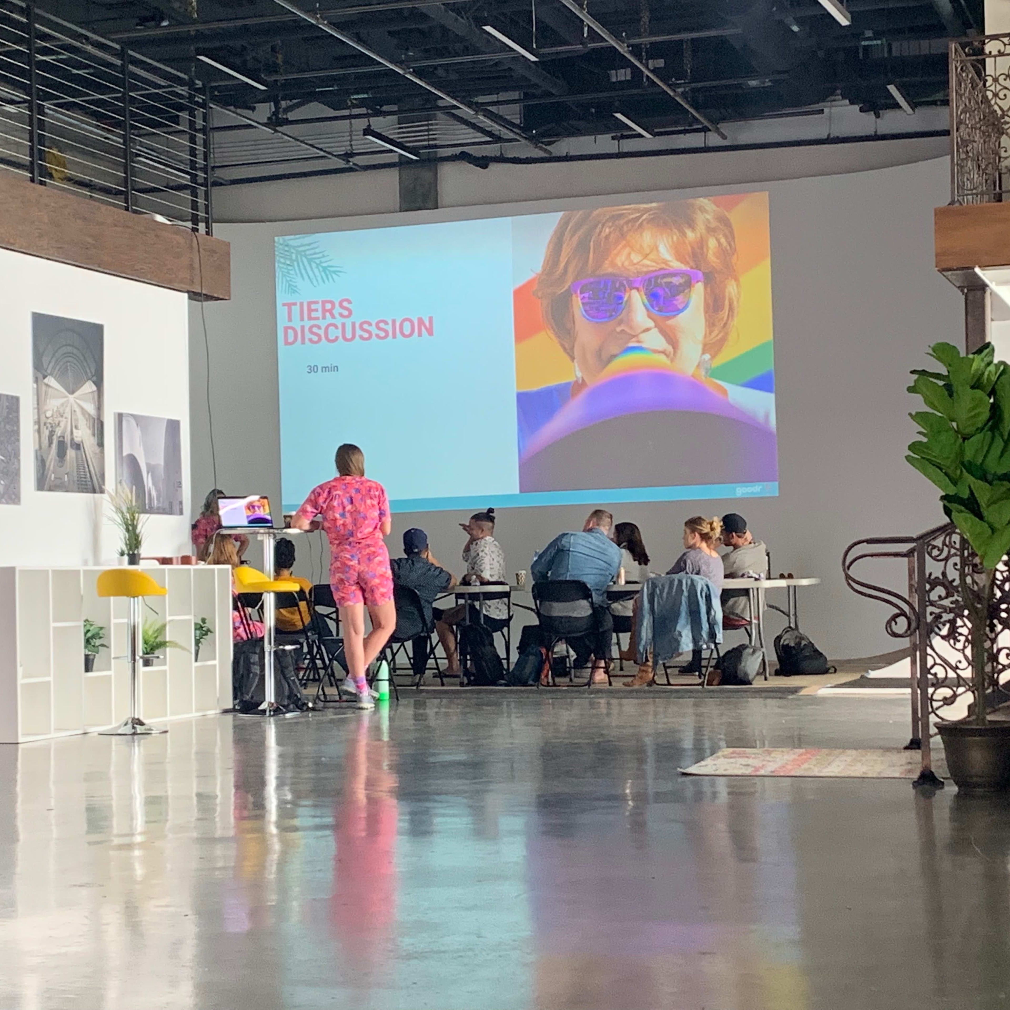 A group of people sits at a table in a well-lit room, with a large screen displaying the words "TIERS DISCUSSION" alongside a colorful image.