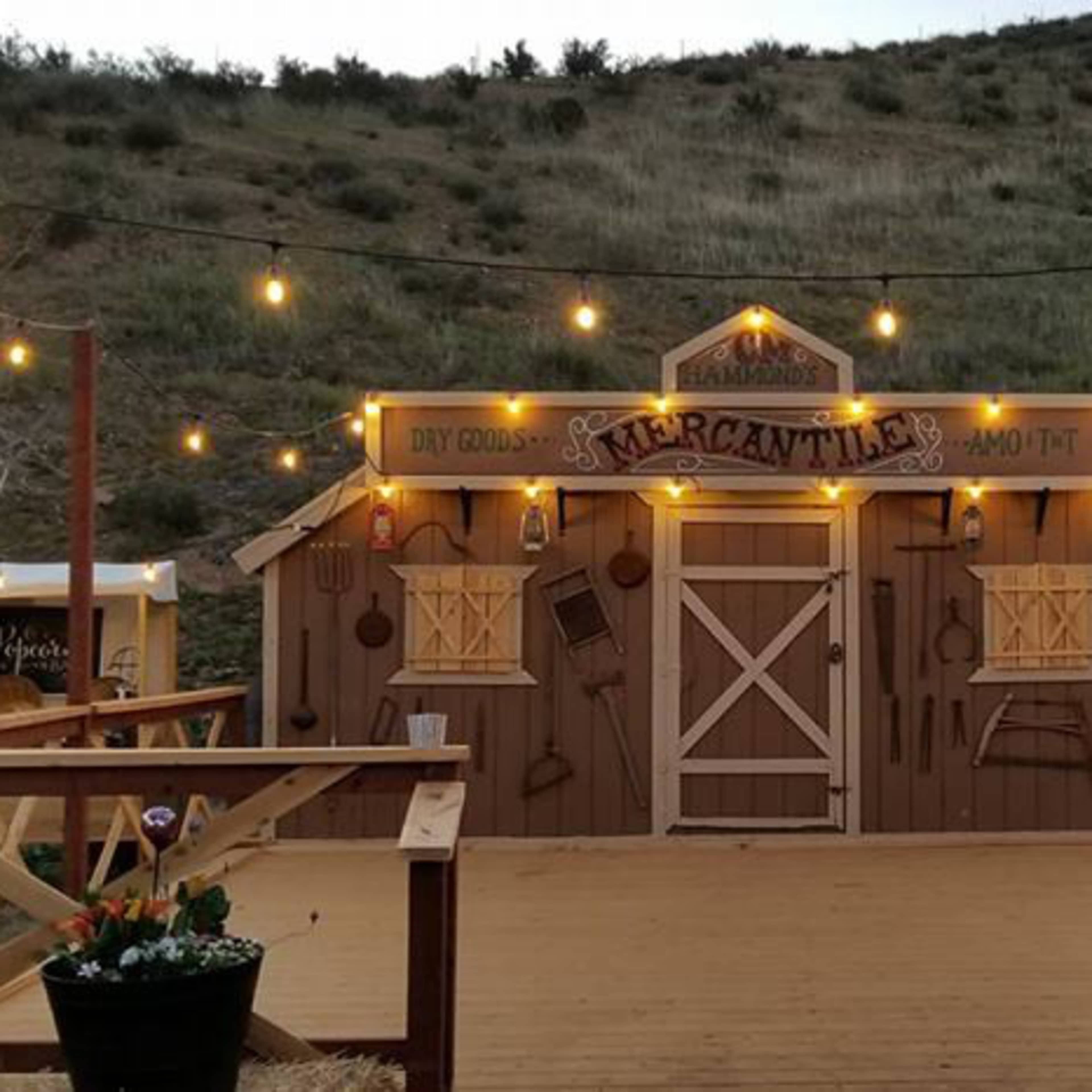 A rustic building with a wooden facade, illuminated by string lights, situated in a rural outdoor setting with a hillside in the background.