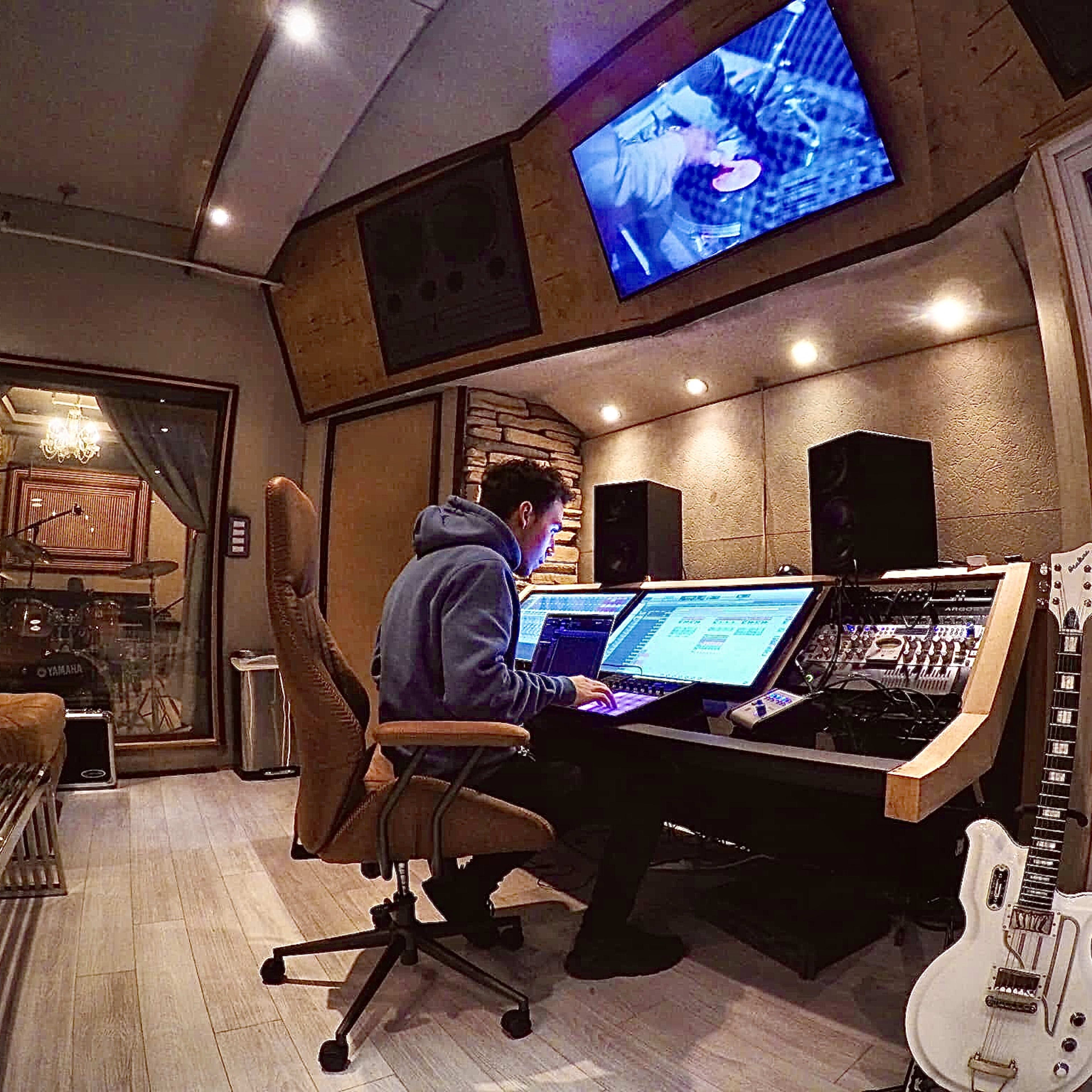 A person sits at a large audio mixing console in a sound studio, working on a computer with monitors displaying music software.