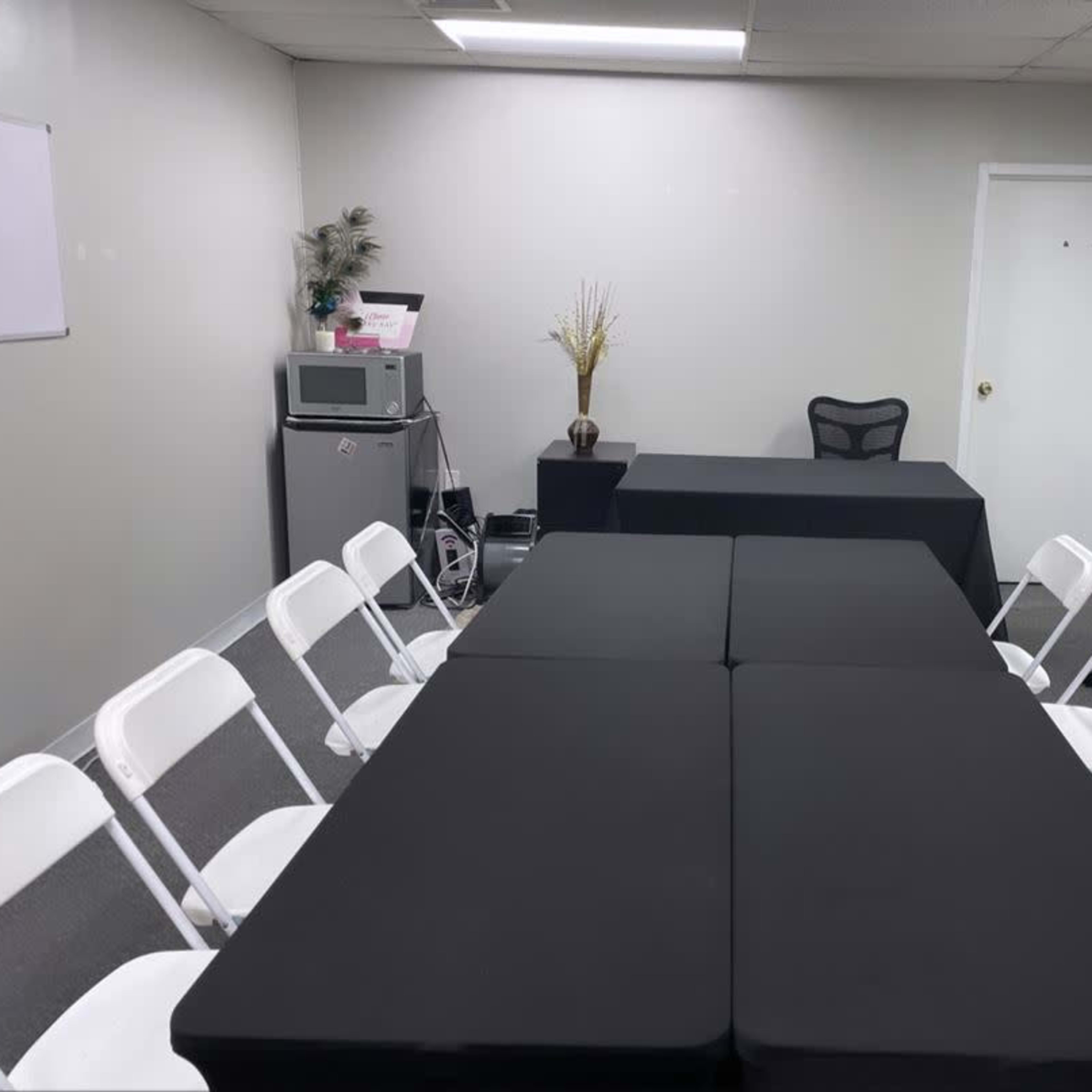 A meeting room features two black tables arranged in a U-shape, surrounded by white folding chairs, with a small podium and a microwave in the corner.