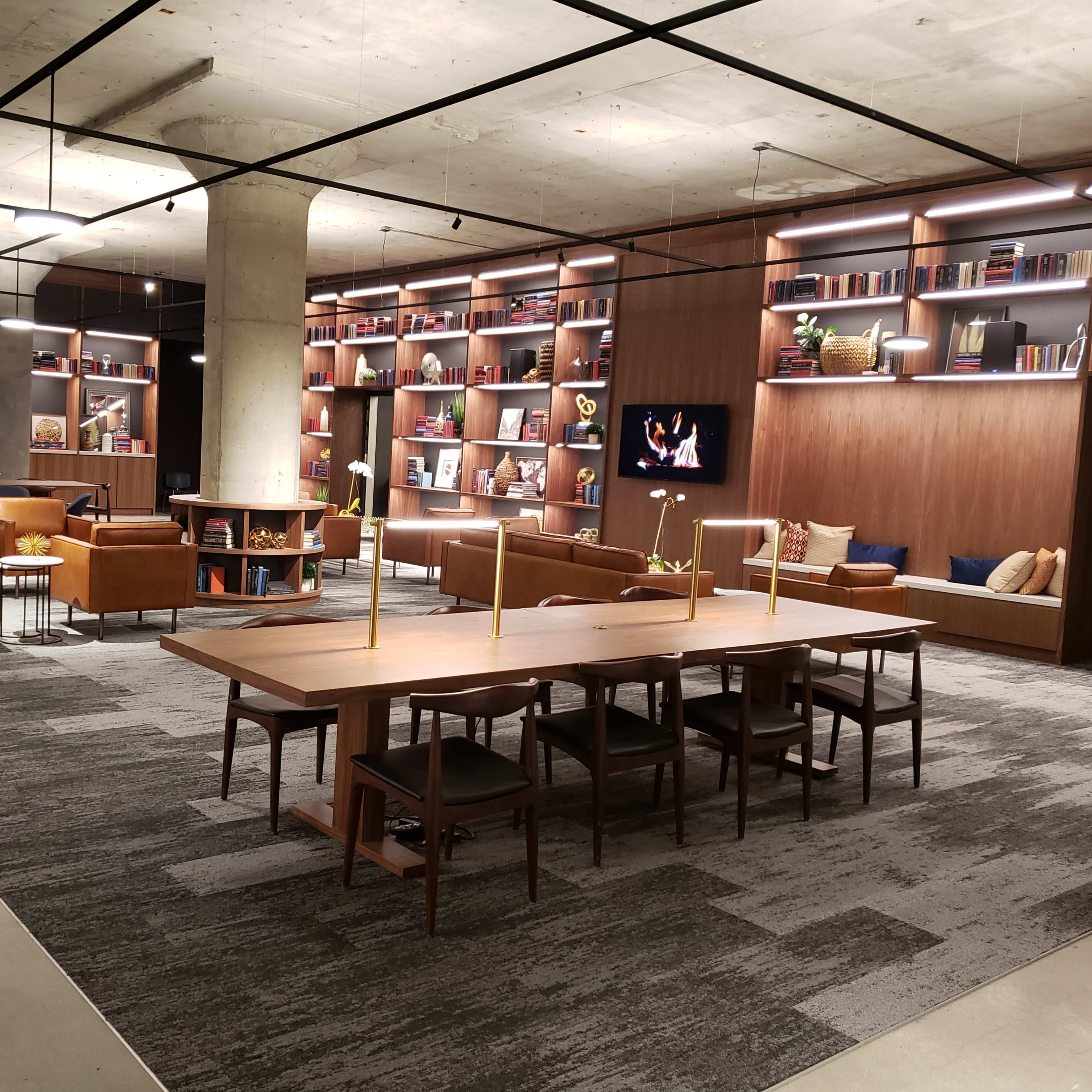A modern library or lounge area with wooden furniture, shelves filled with books, and soft seating arranged around a central table.