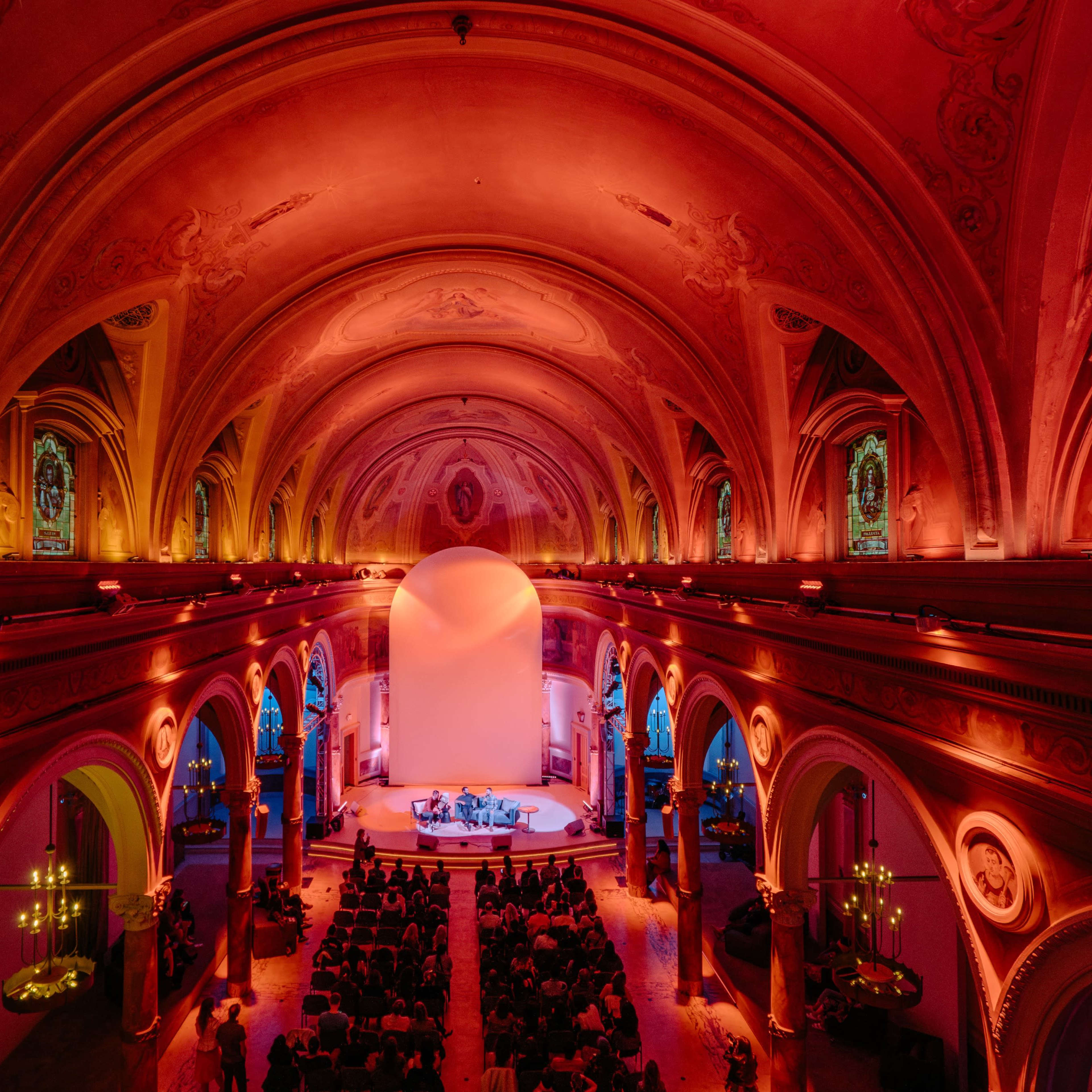 A spacious concert hall characterized by an elaborately arched ceiling and a stage set in an illuminated area with an audience seated below.