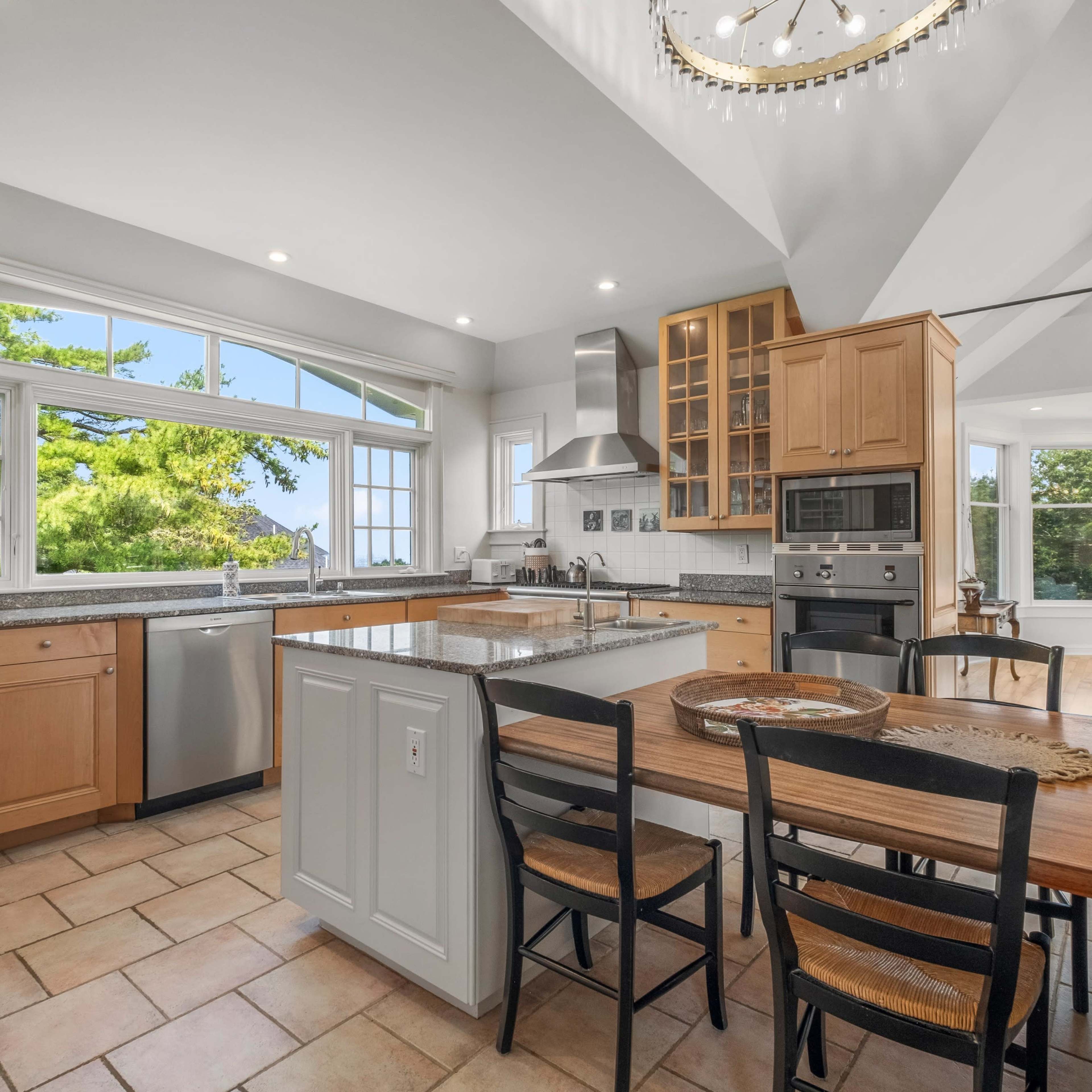 A modern kitchen features wooden cabinets, a central island, and large windows overlooking a green landscape.
