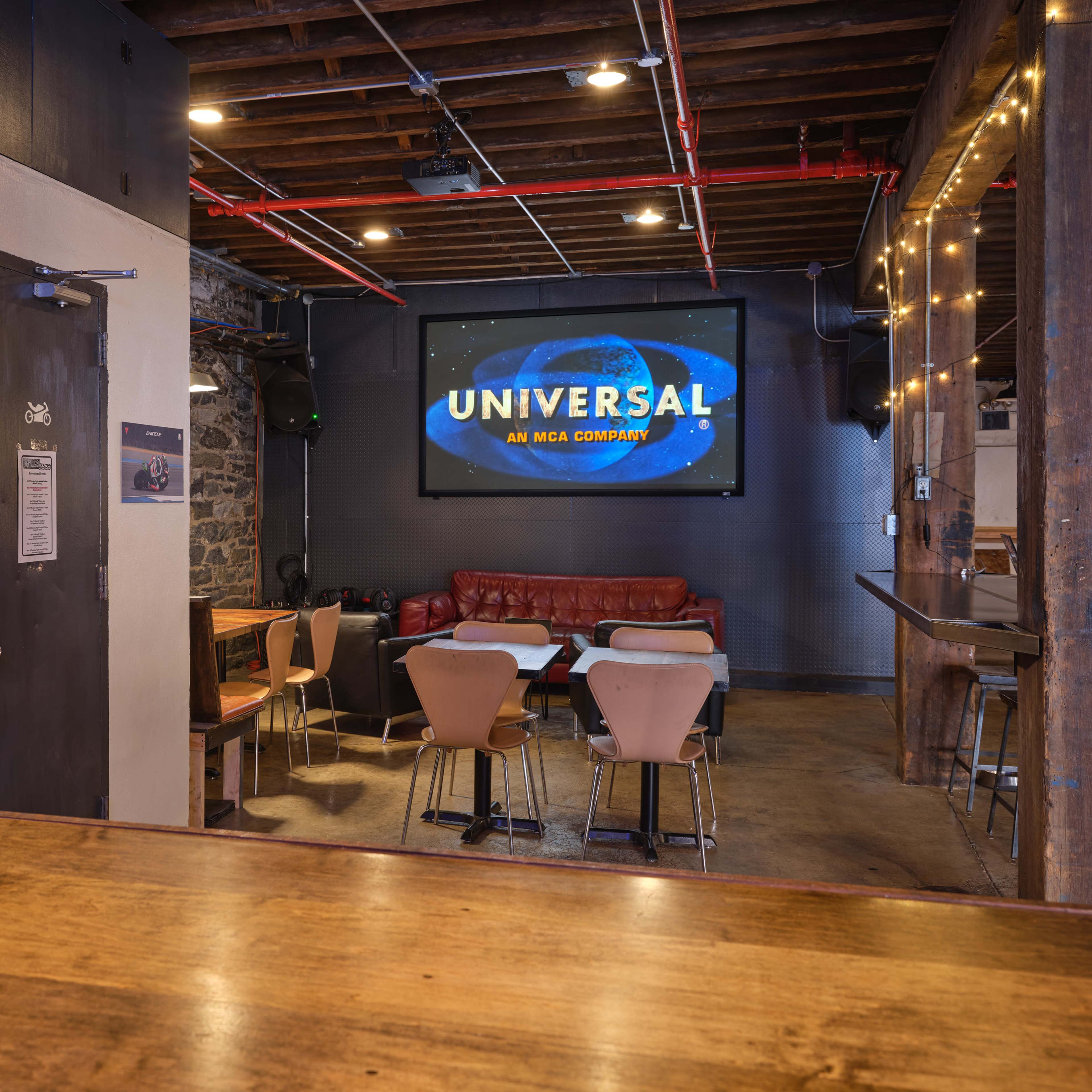 A dimly lit bar area features a large screen displaying "UNIVERSAL" on one wall, with seating arranged around tables and a leather couch along the opposite wall.