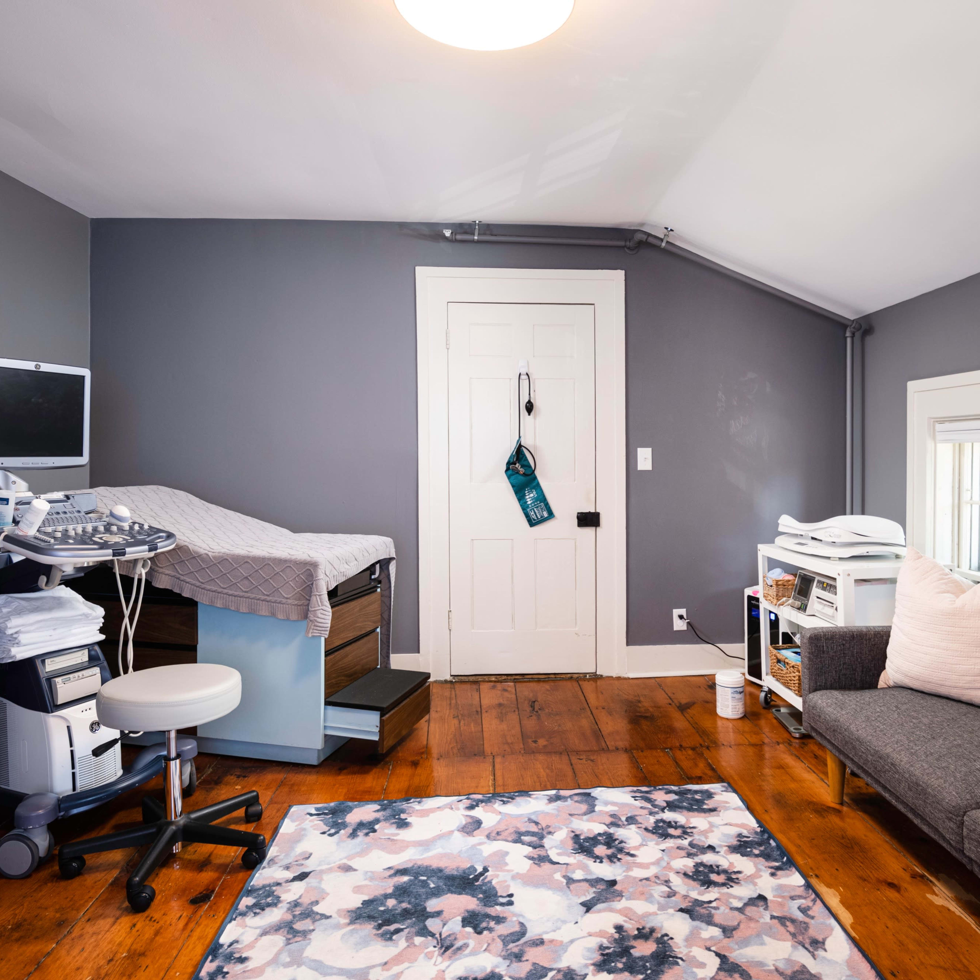 A small room with a medical examination table, a desk with medical equipment, and a sofa, featuring wooden floors and a window.
