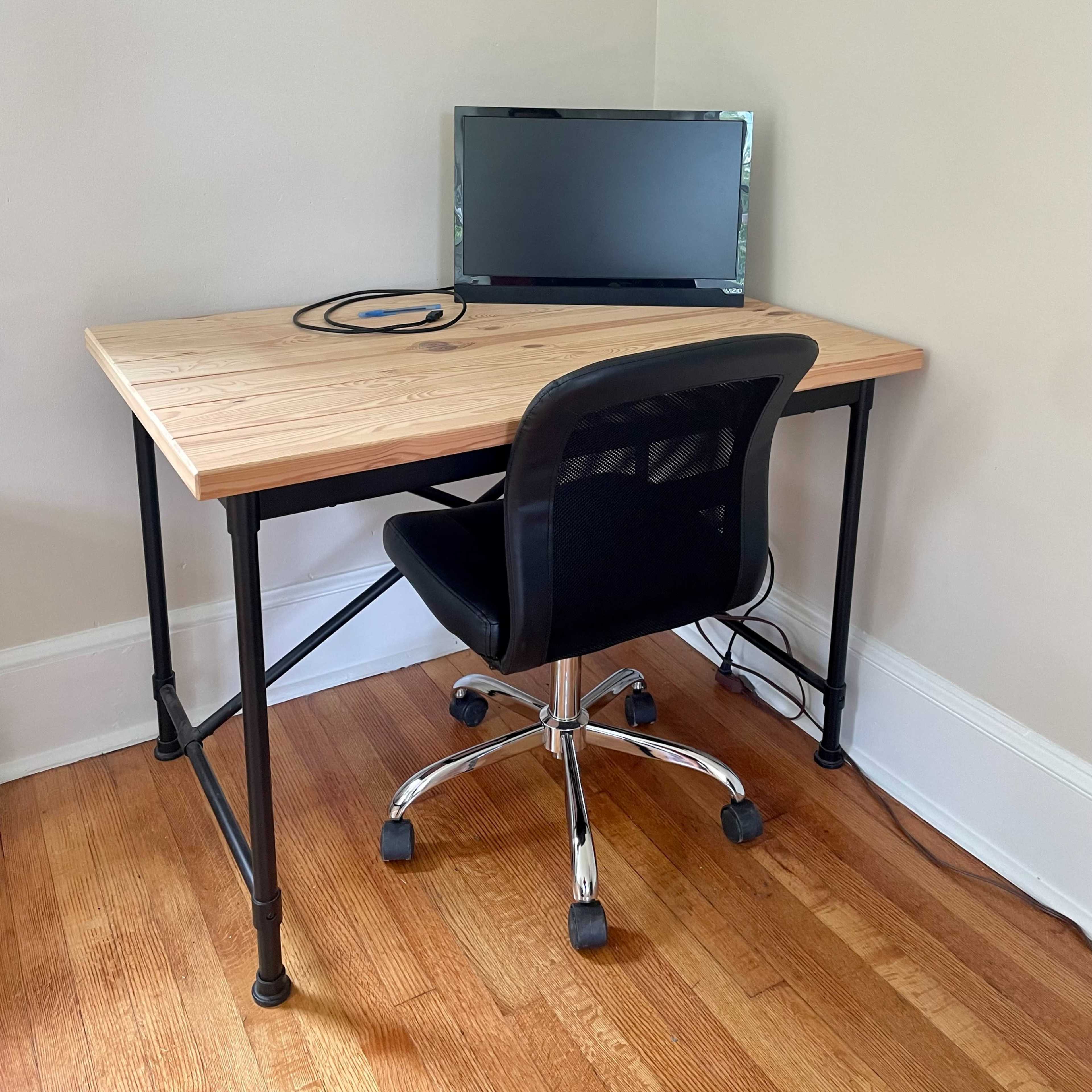 A wooden desk with a black office chair and a monitor is positioned in a corner of a room with light-colored walls and hardwood flooring.