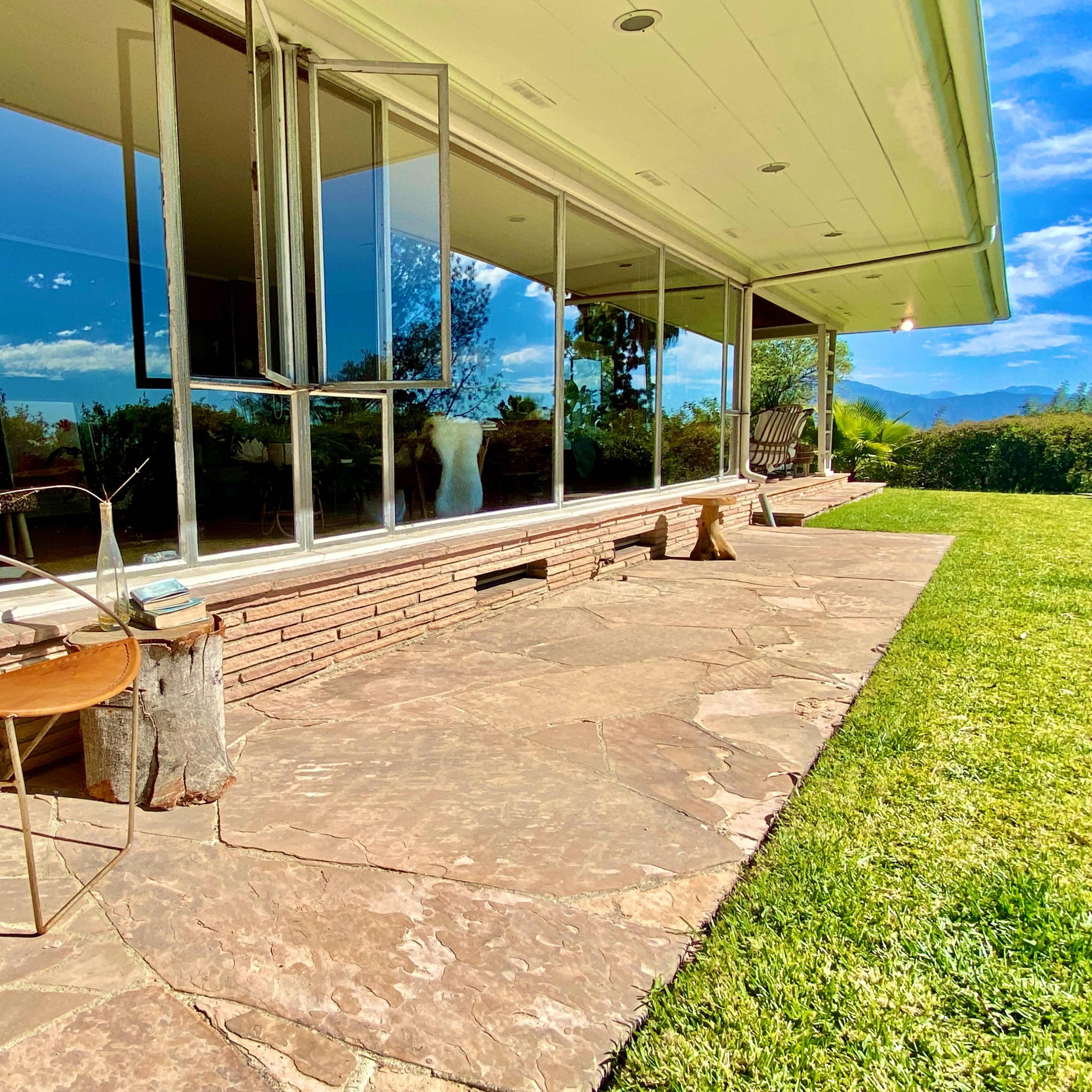 A modern home features a stone patio with a metal chair and large glass windows overlooking a grassy landscape.