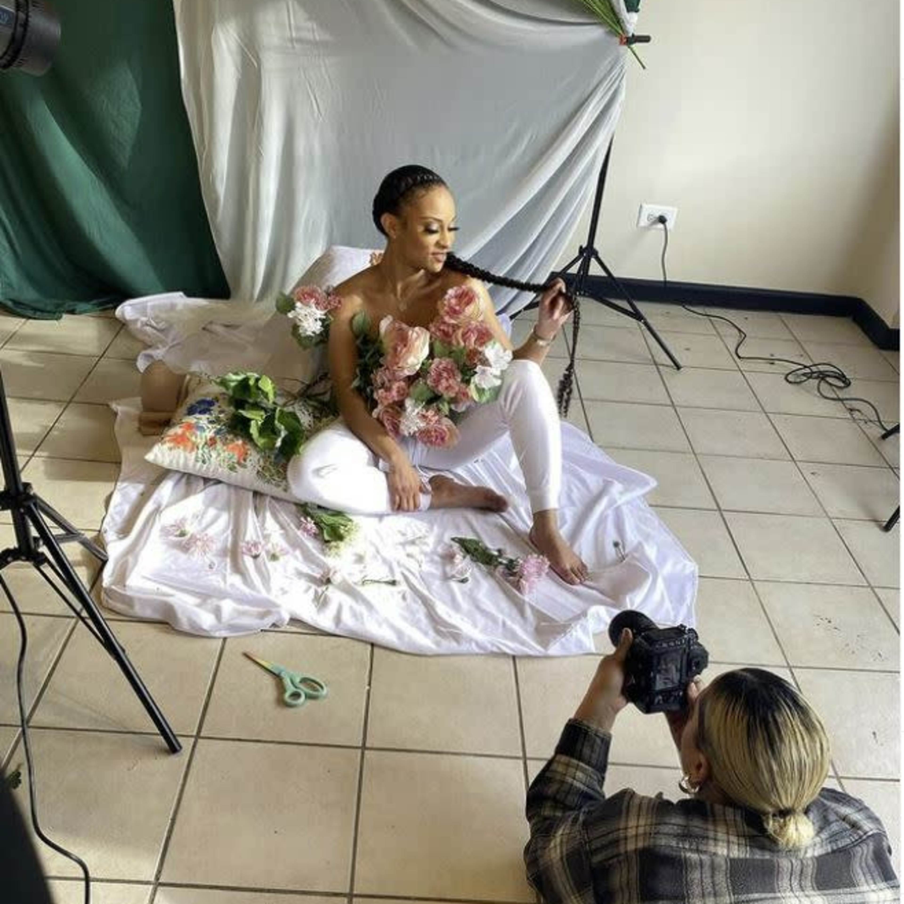 A model poses with flowers in her arms while a photographer captures the scene on a tiled floor in a studio setting.