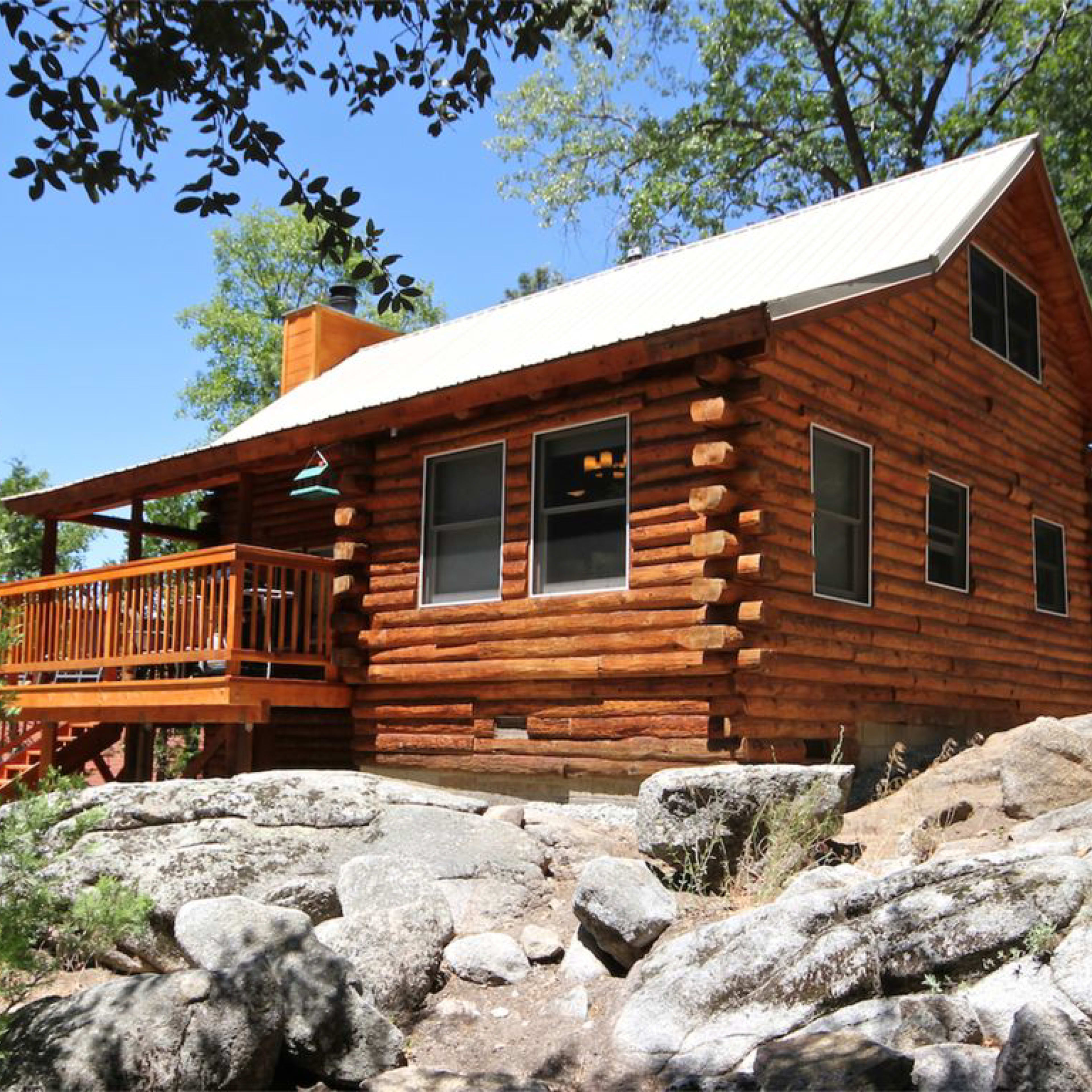 A wooden log cabin with a metal roof is set among large rocks and trees under a clear blue sky.