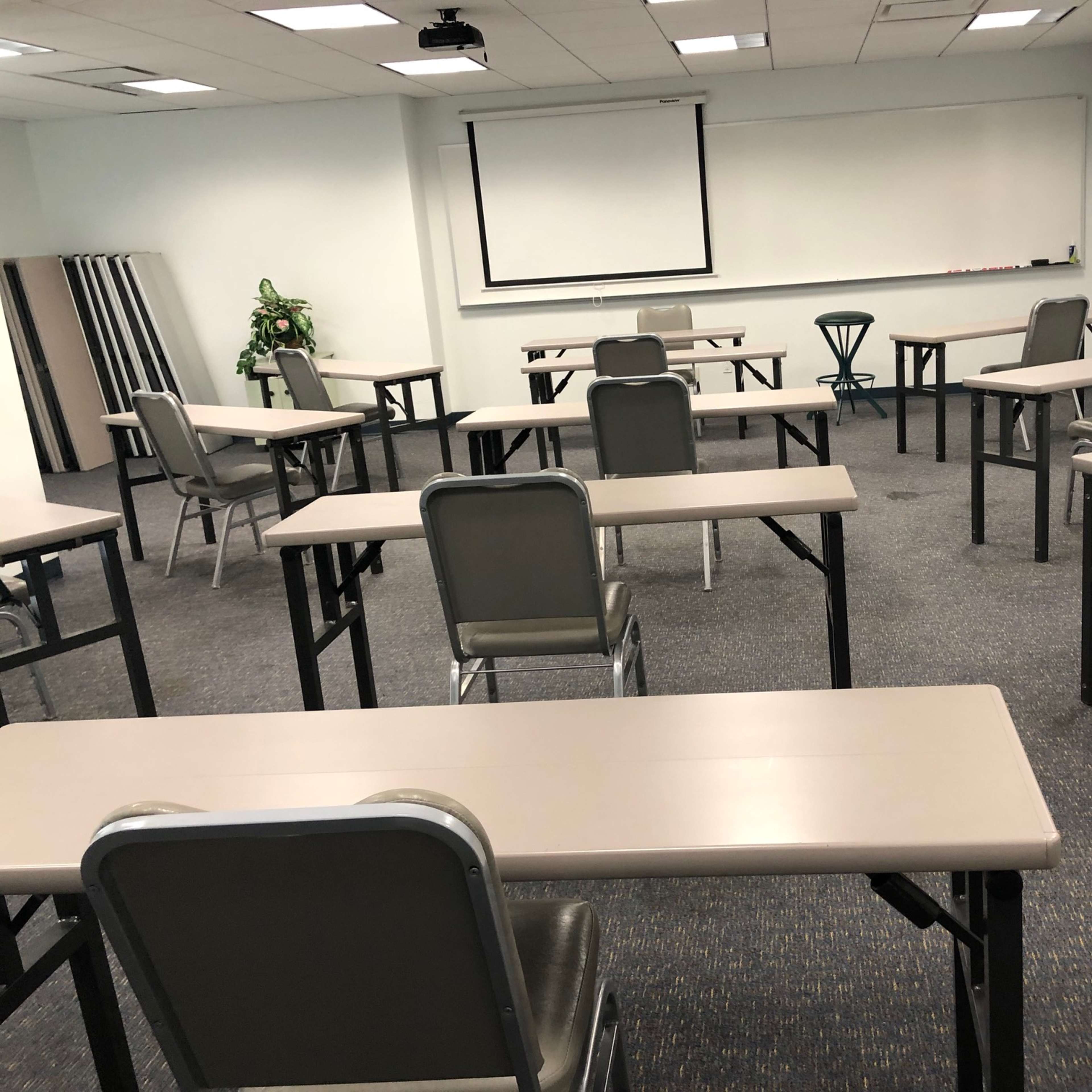 A sparsely arranged classroom with several tables and chairs facing a blank whiteboard at the front.
