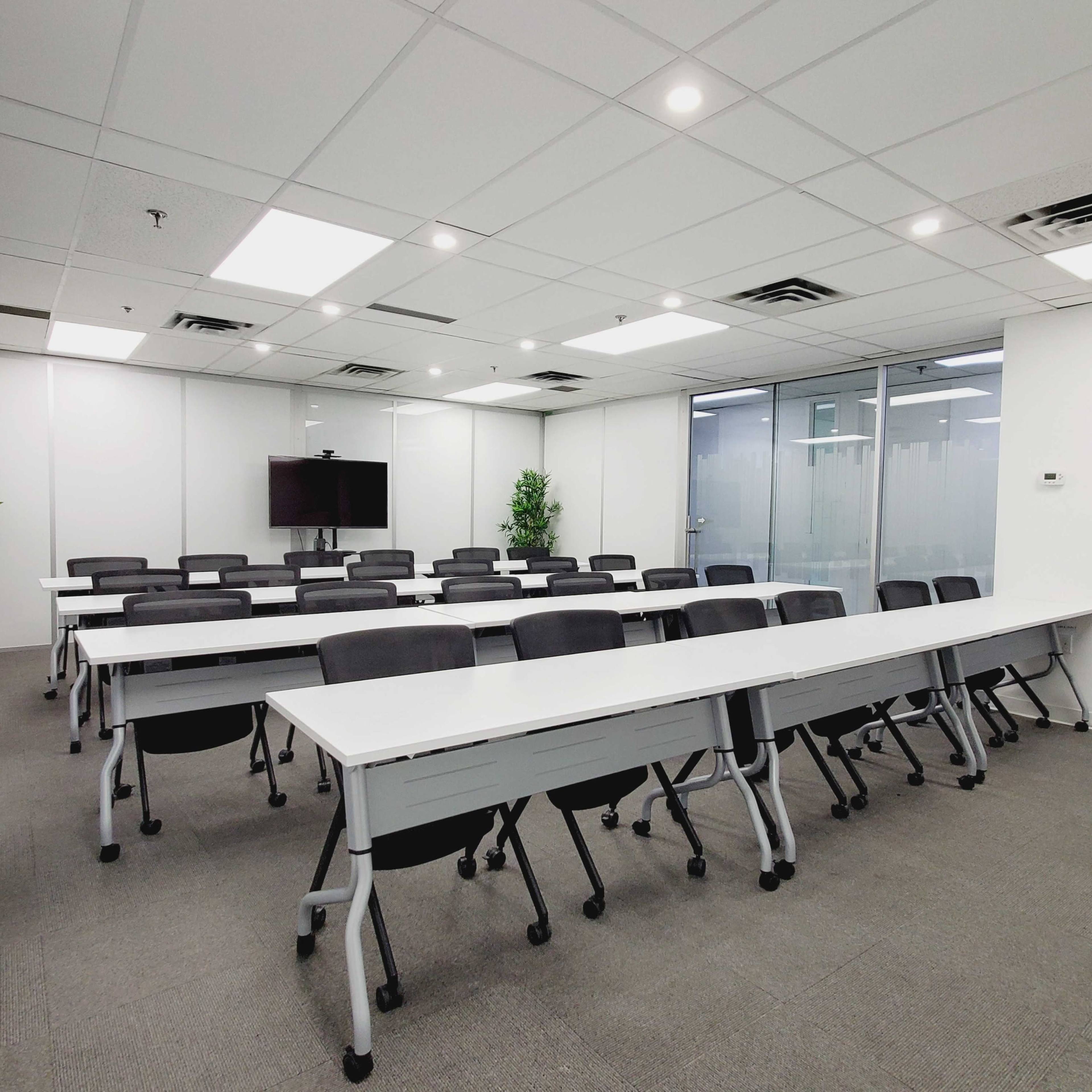 A modern conference room with rows of tables and chairs arranged neatly, featuring a large screen at one end and large windows allowing natural light.