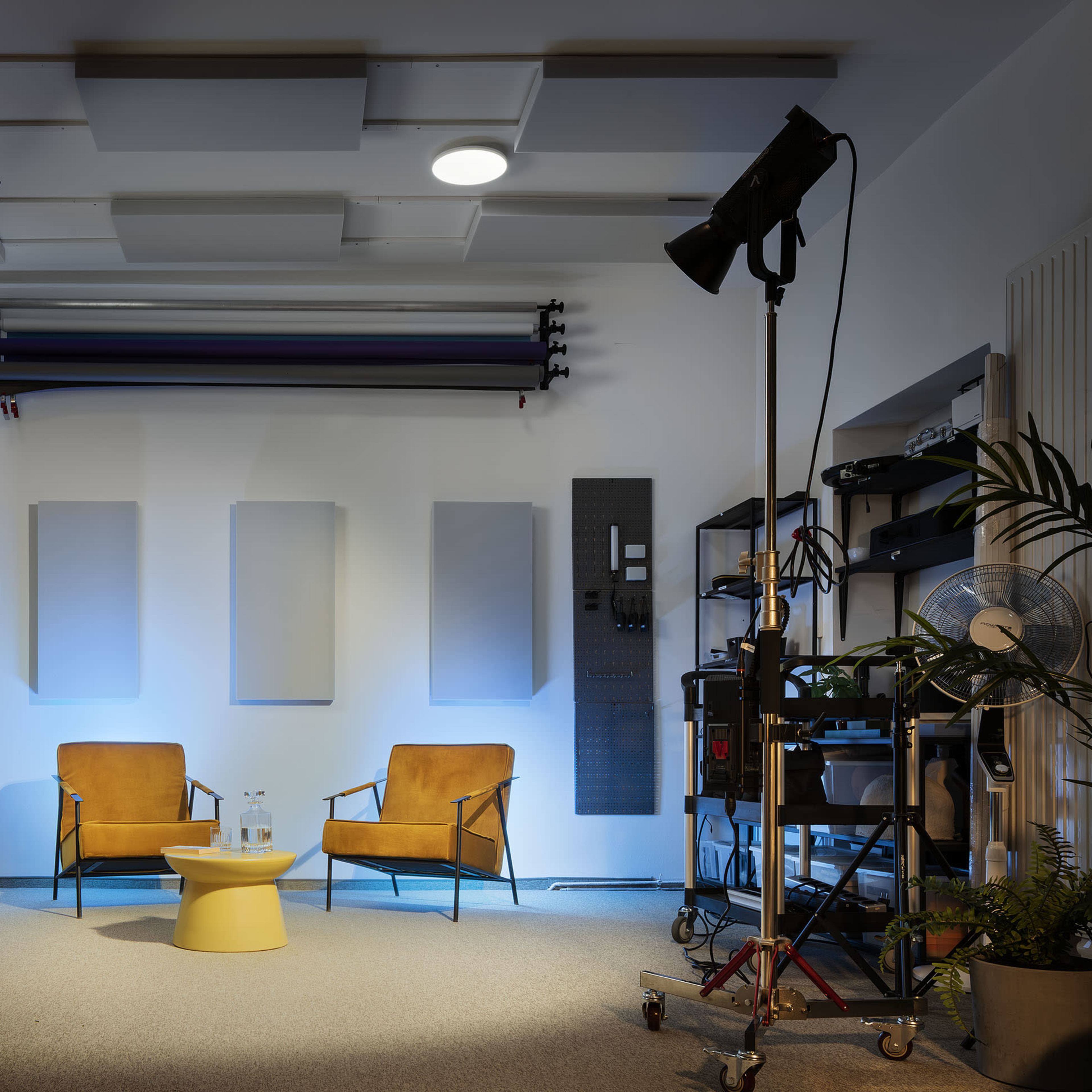 A well-lit studio space featuring two yellow chairs facing a small round table, with studio equipment and sound-absorbing panels in the background.