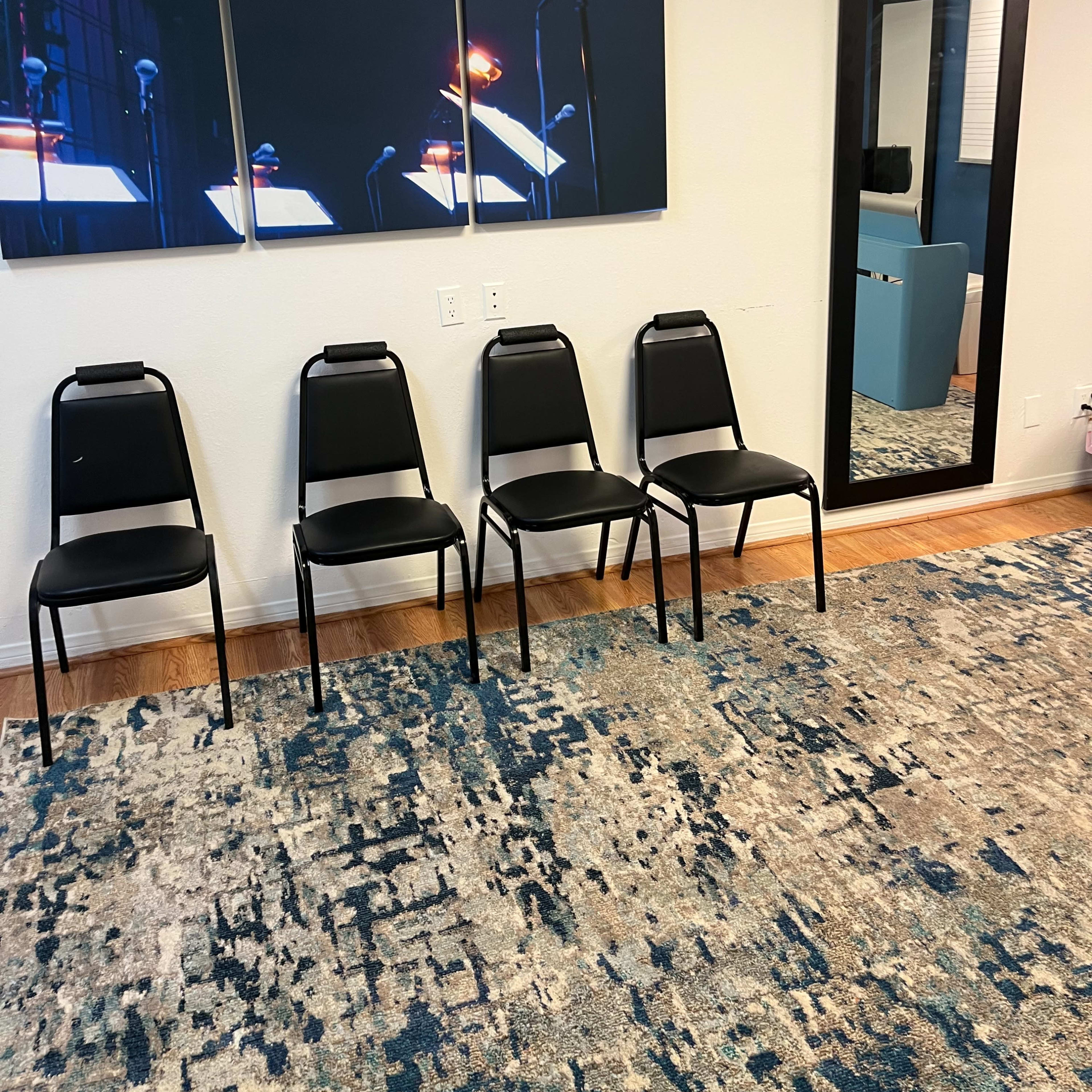 A row of four black chairs lined up against a wall with three framed pictures above them and a large mirror adjacent to the chairs.