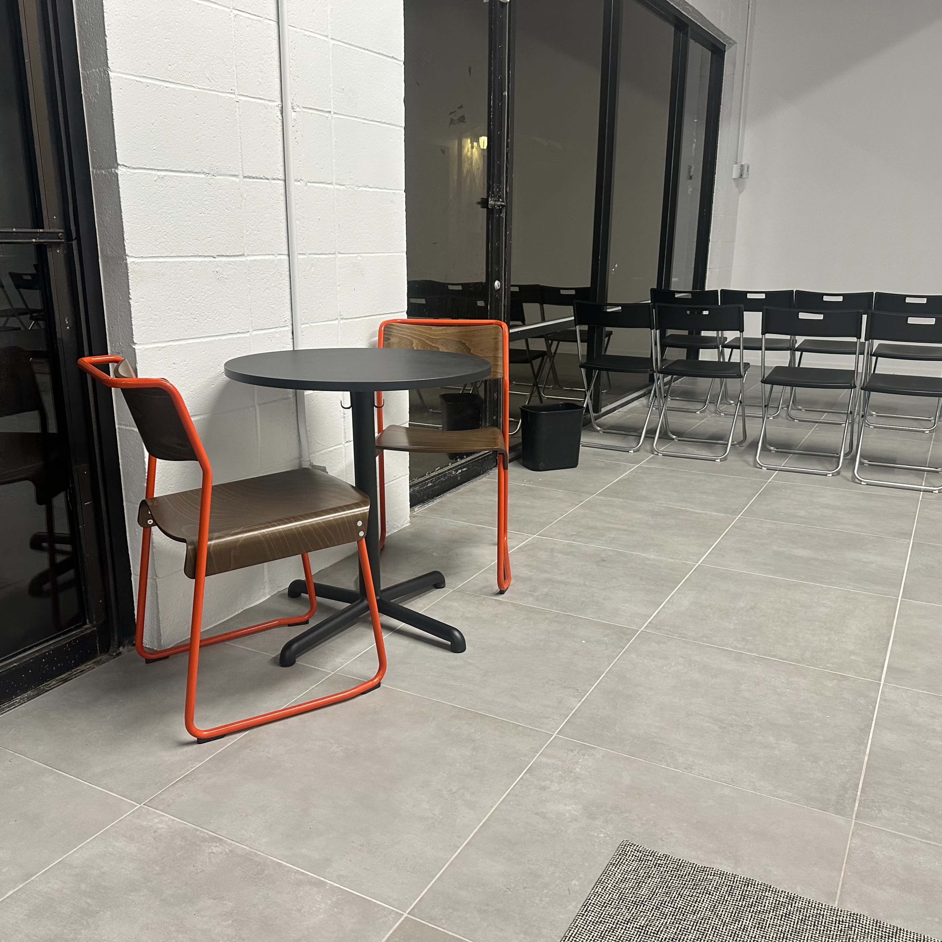 A small table with two chairs is placed near a glass wall in a sparsely furnished room featuring additional stacked chairs in the background.