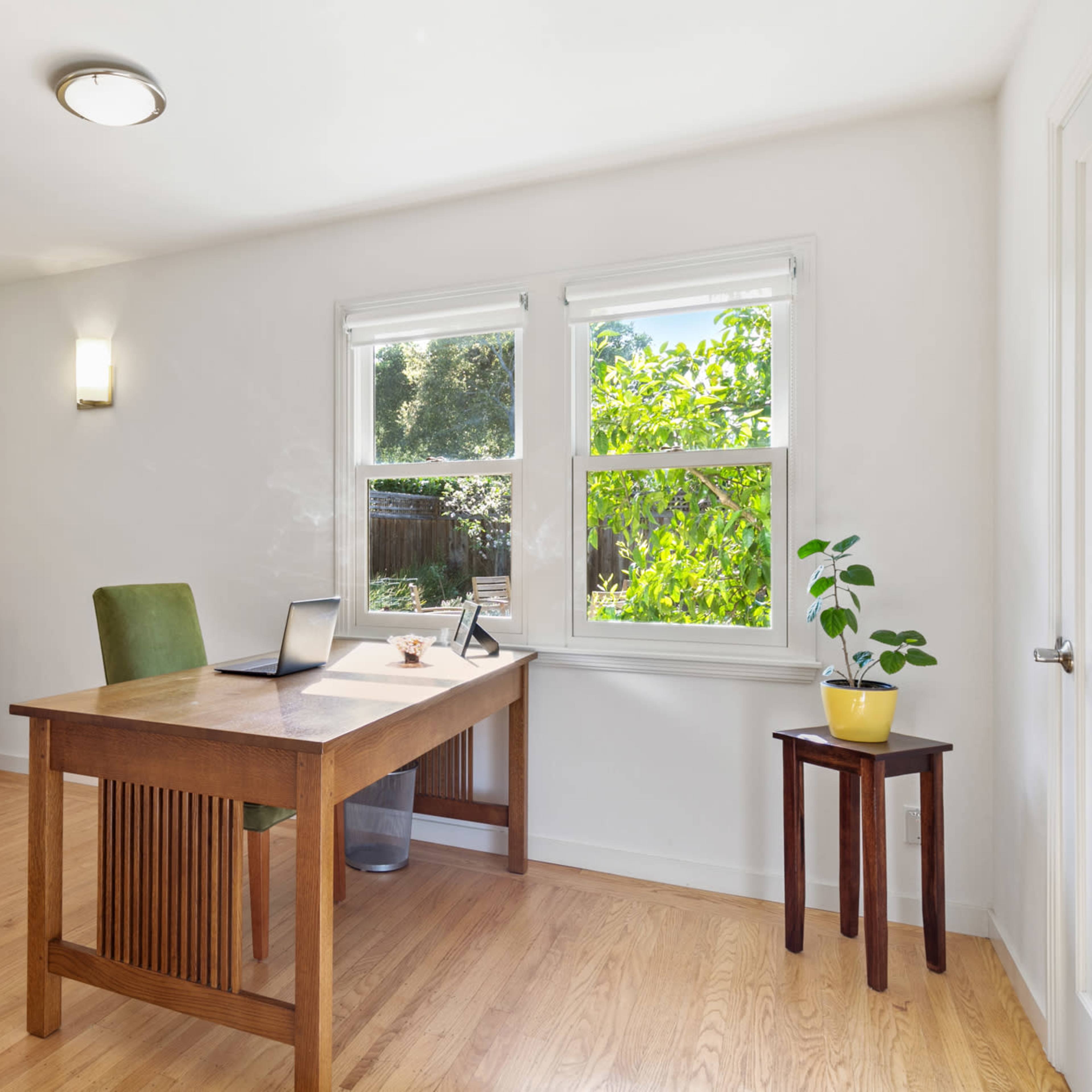 A bright home office features a wooden desk, a green chair, shelves with glass doors, and windows overlooking a garden.