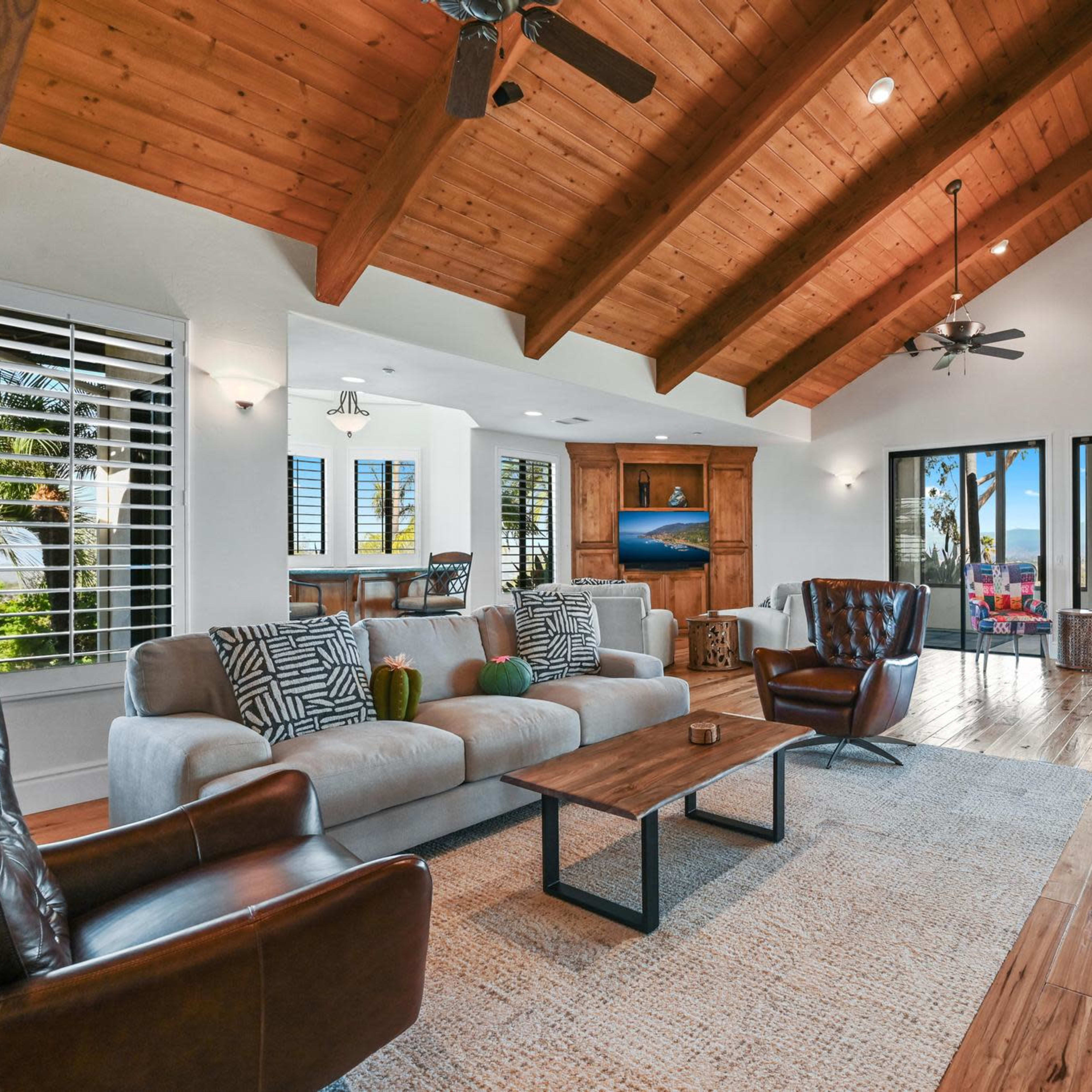 A spacious living room with exposed wooden beams, a stone fireplace, and large windows opening to a view of the outdoors.