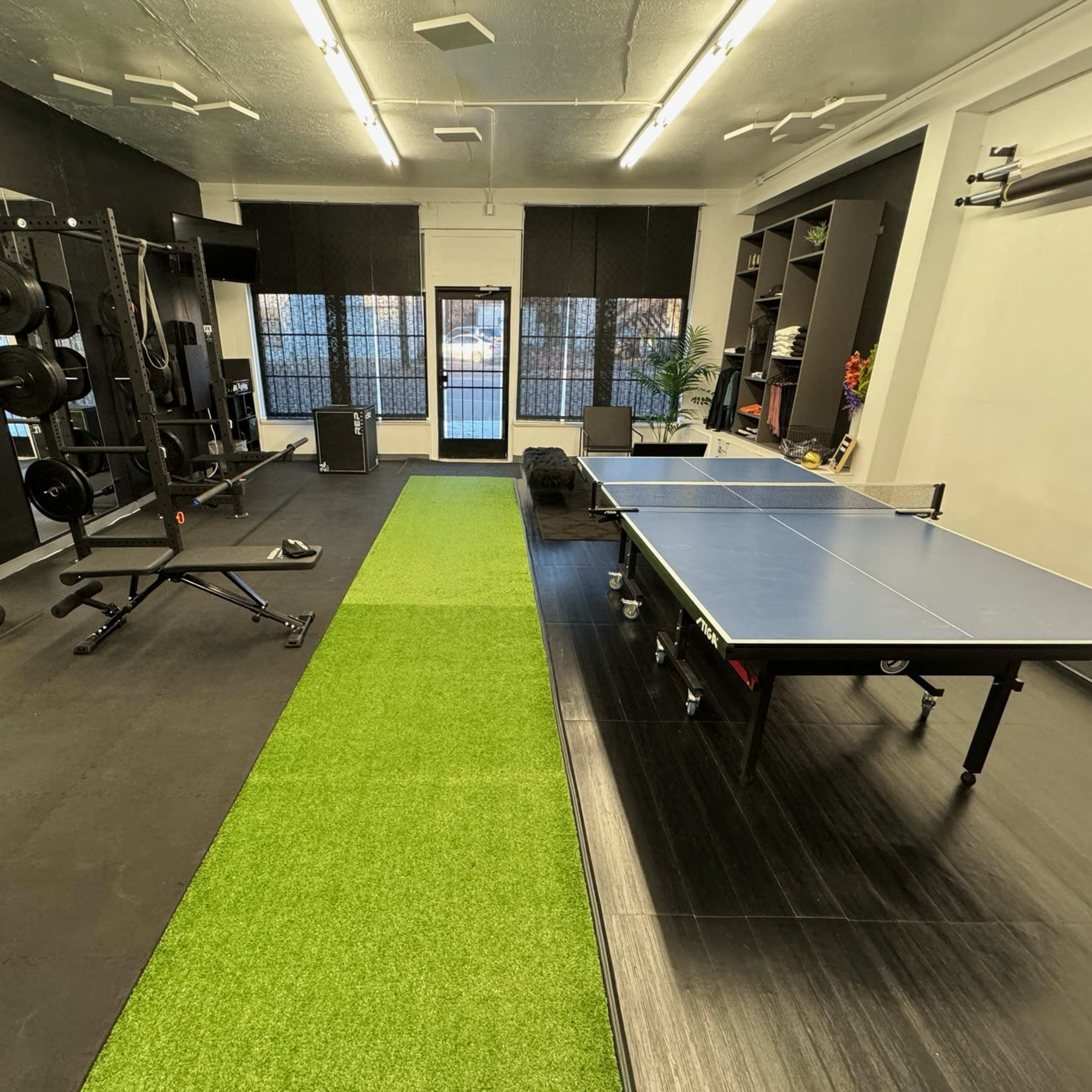 The room features a gym area with exercise equipment on one side and a ping pong table on the other, separated by an area of artificial grass.