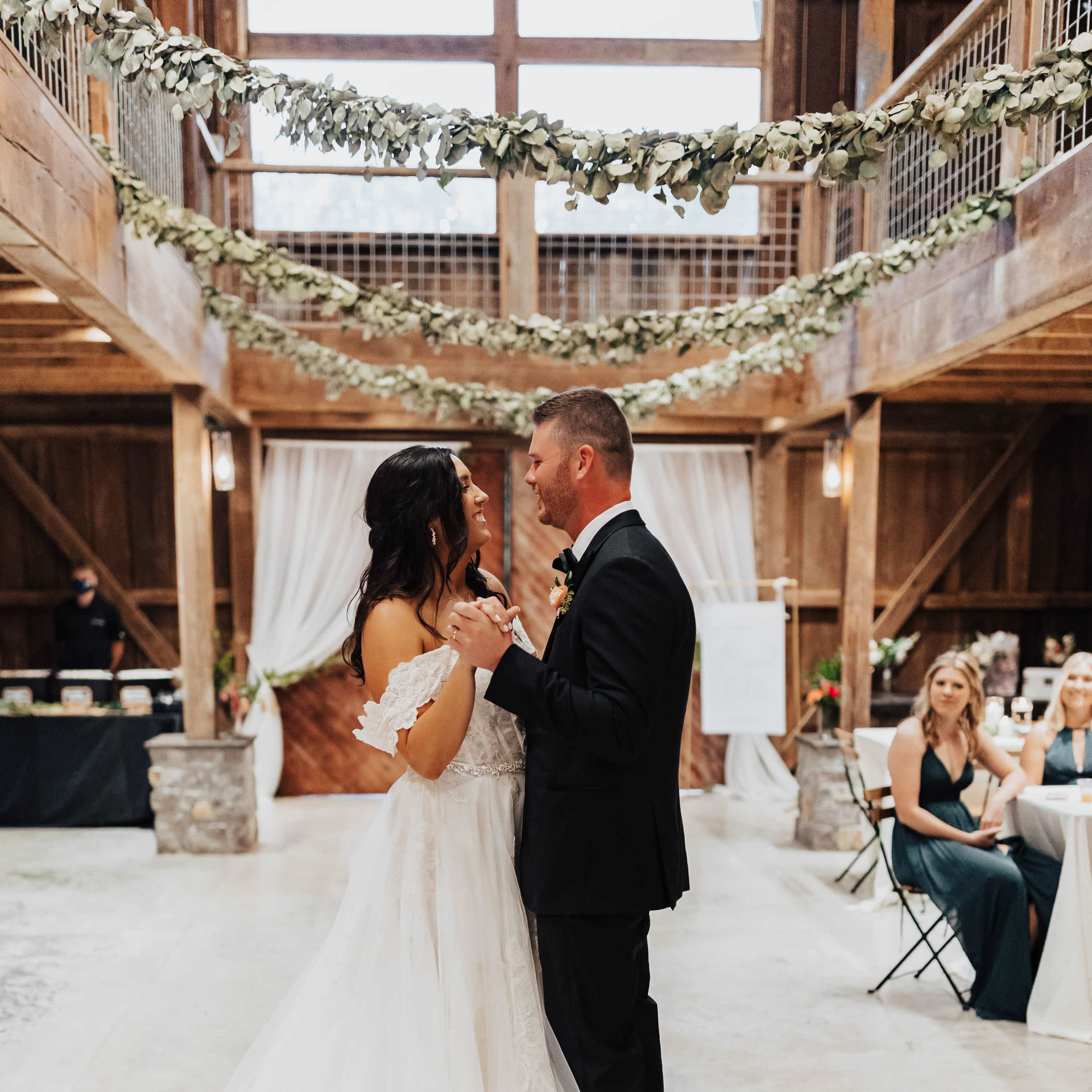 A couple dances together in a rustic barn setting adorned with greenery and floral decorations, while guests watch from seated arrangements.