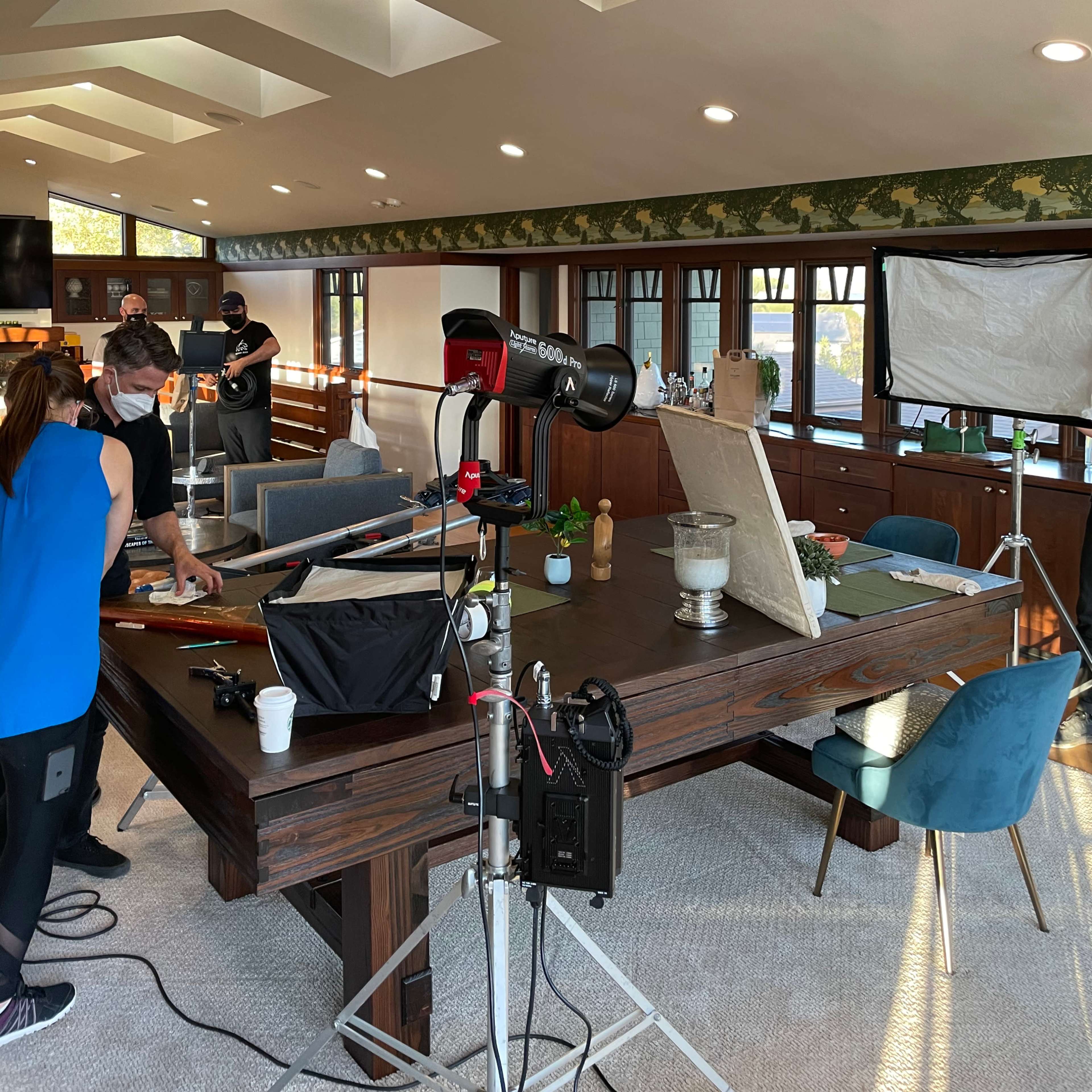 A group of people is preparing a photo shoot in a brightly lit, modern living room with a large wooden table and various equipment set up around it.