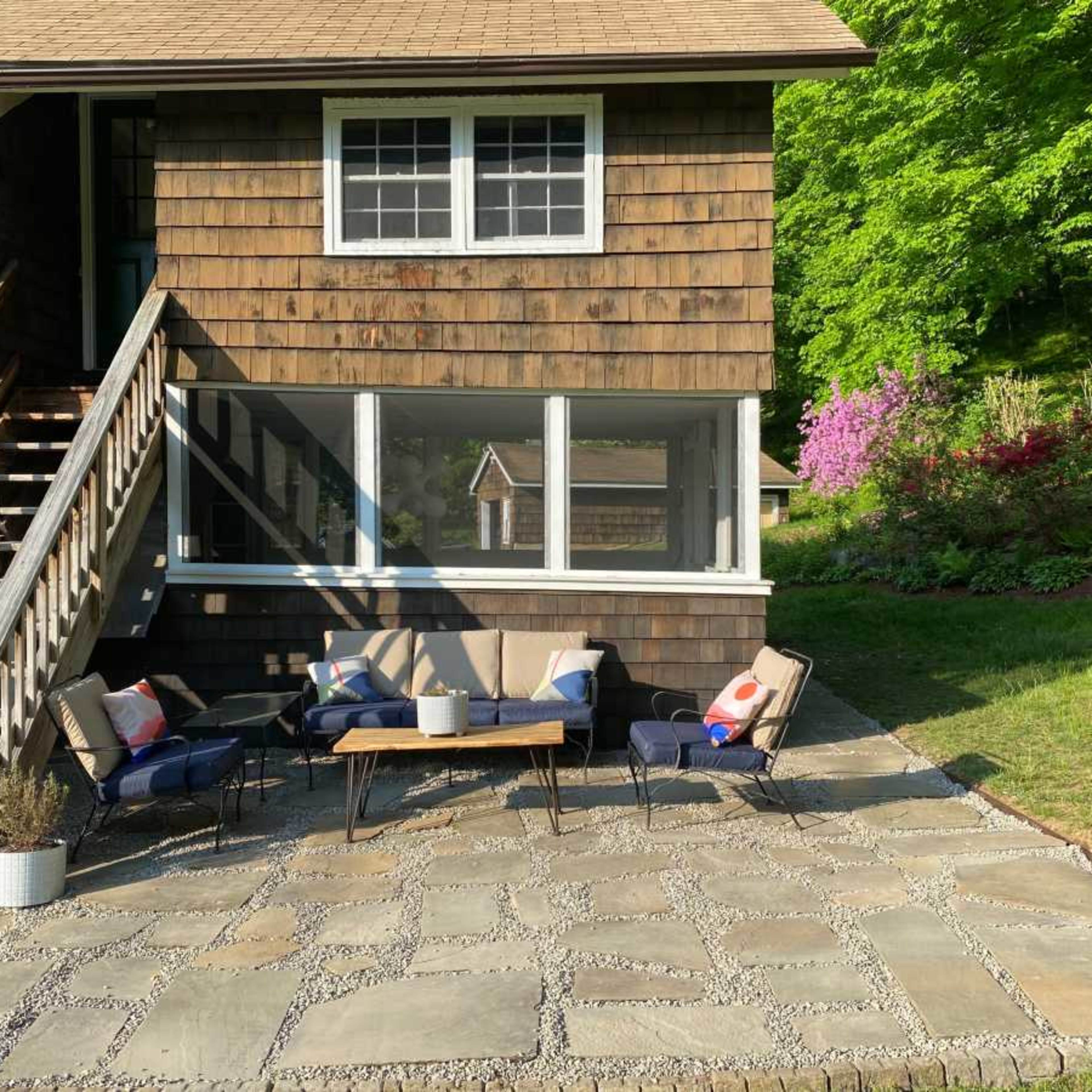 A patio area with a stone floor, featuring a low table surrounded by chairs, is located beside a wooden house and steps leading to the entrance.