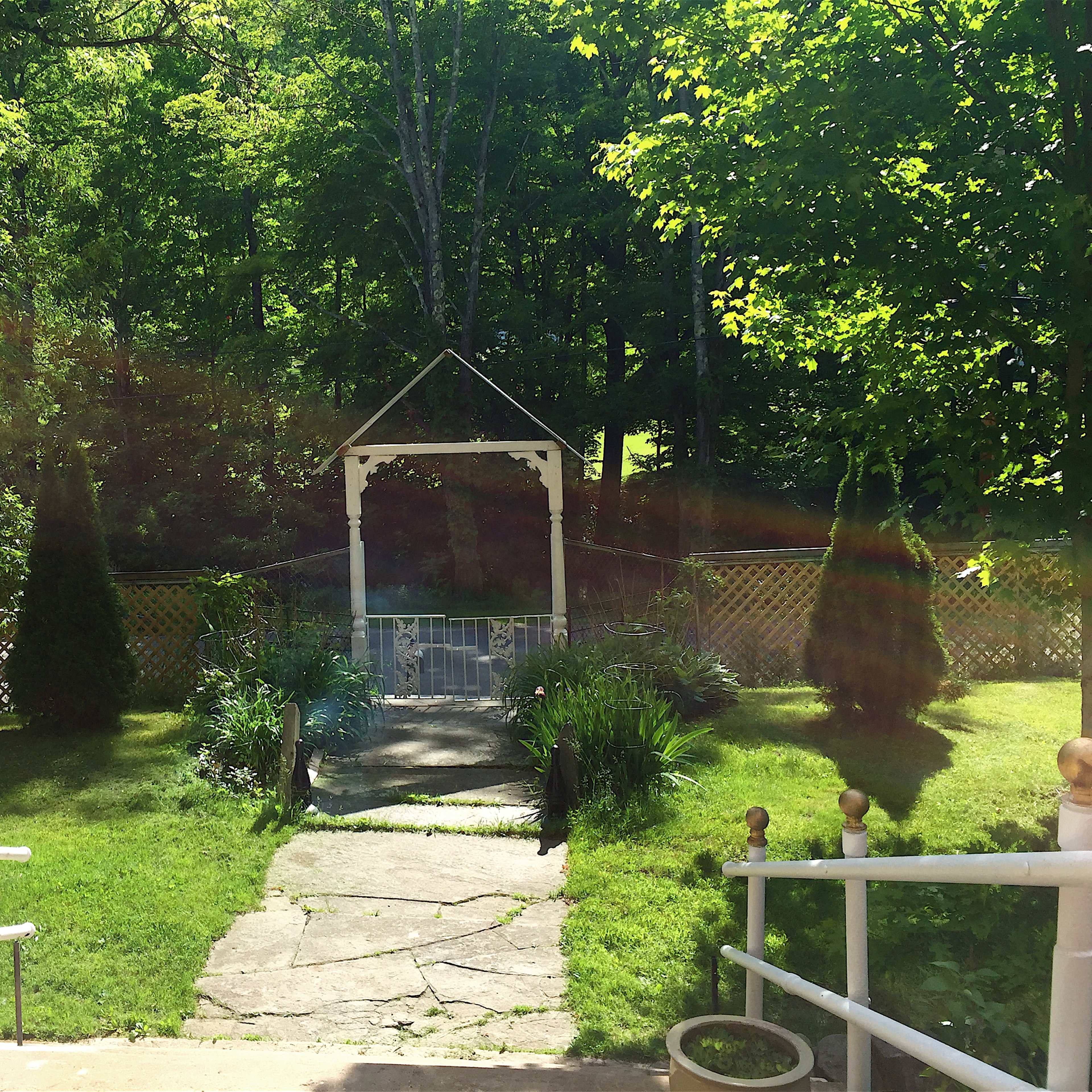 A walkway made of stone leads to a white-gabled gazebo surrounded by green grass and trees.