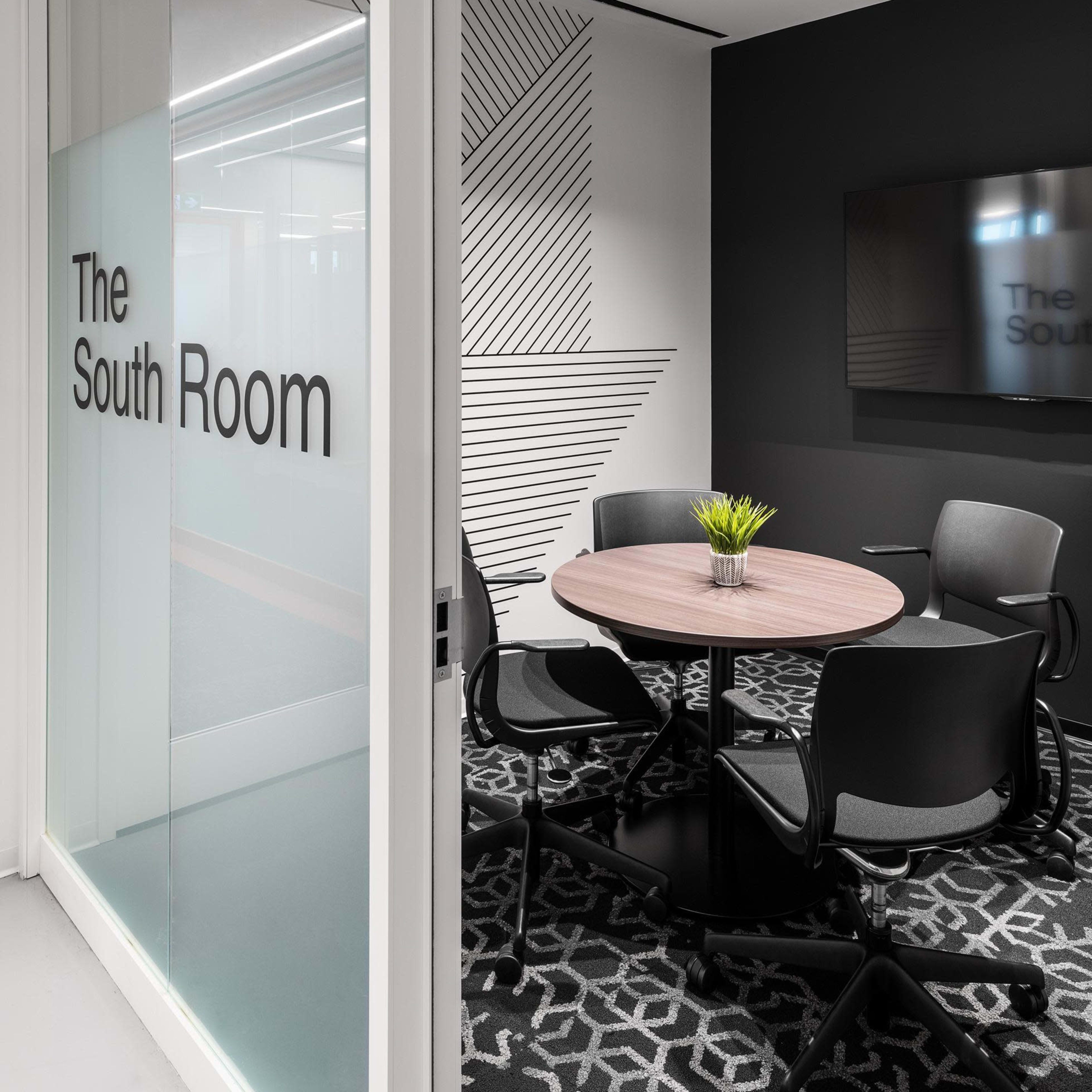 A modern conference room named "The South Room," featuring a round table with four chairs and a large display screen on the wall.