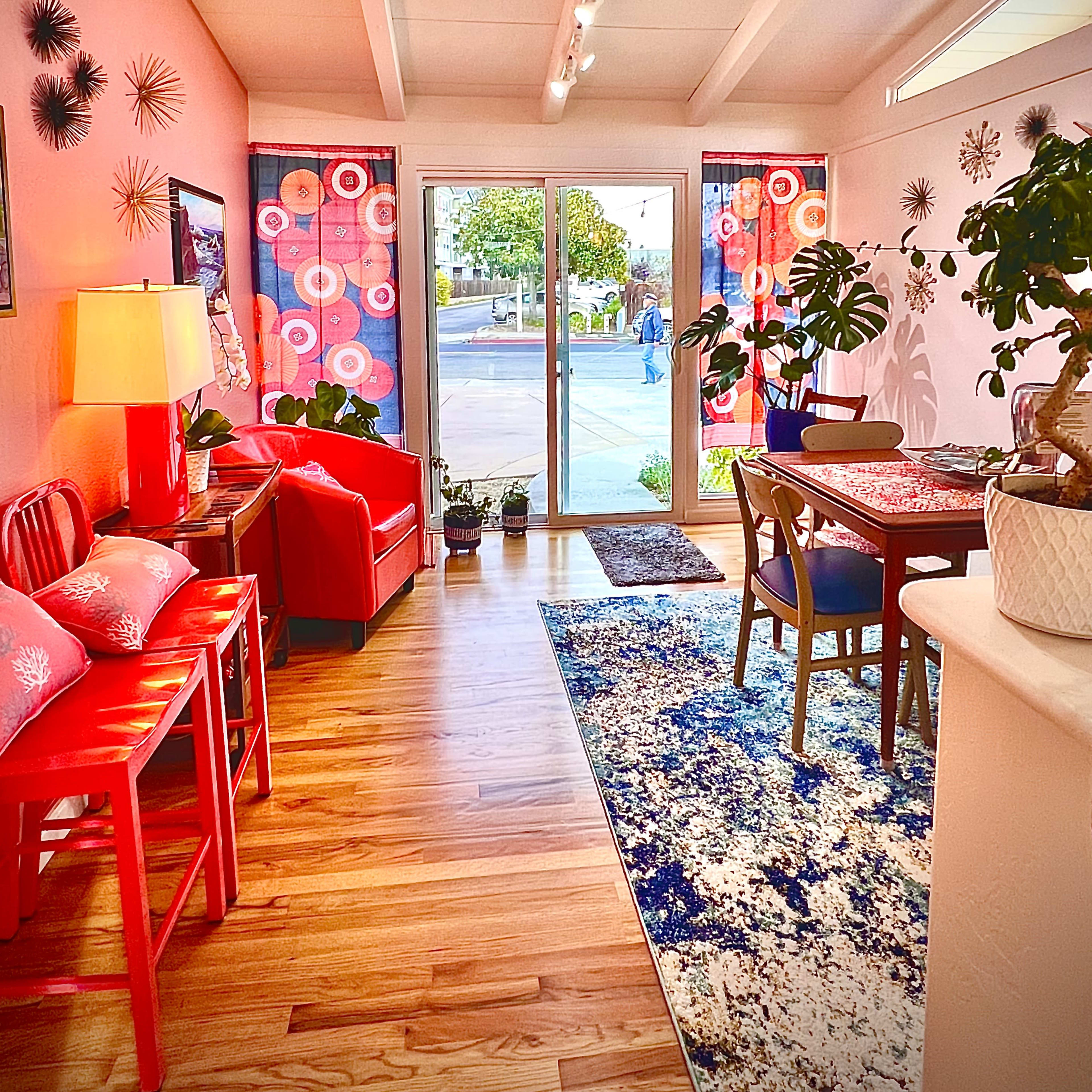 A brightly lit living space featuring a red sofa and chairs, a dining table, and large windows opening to an outdoor view.