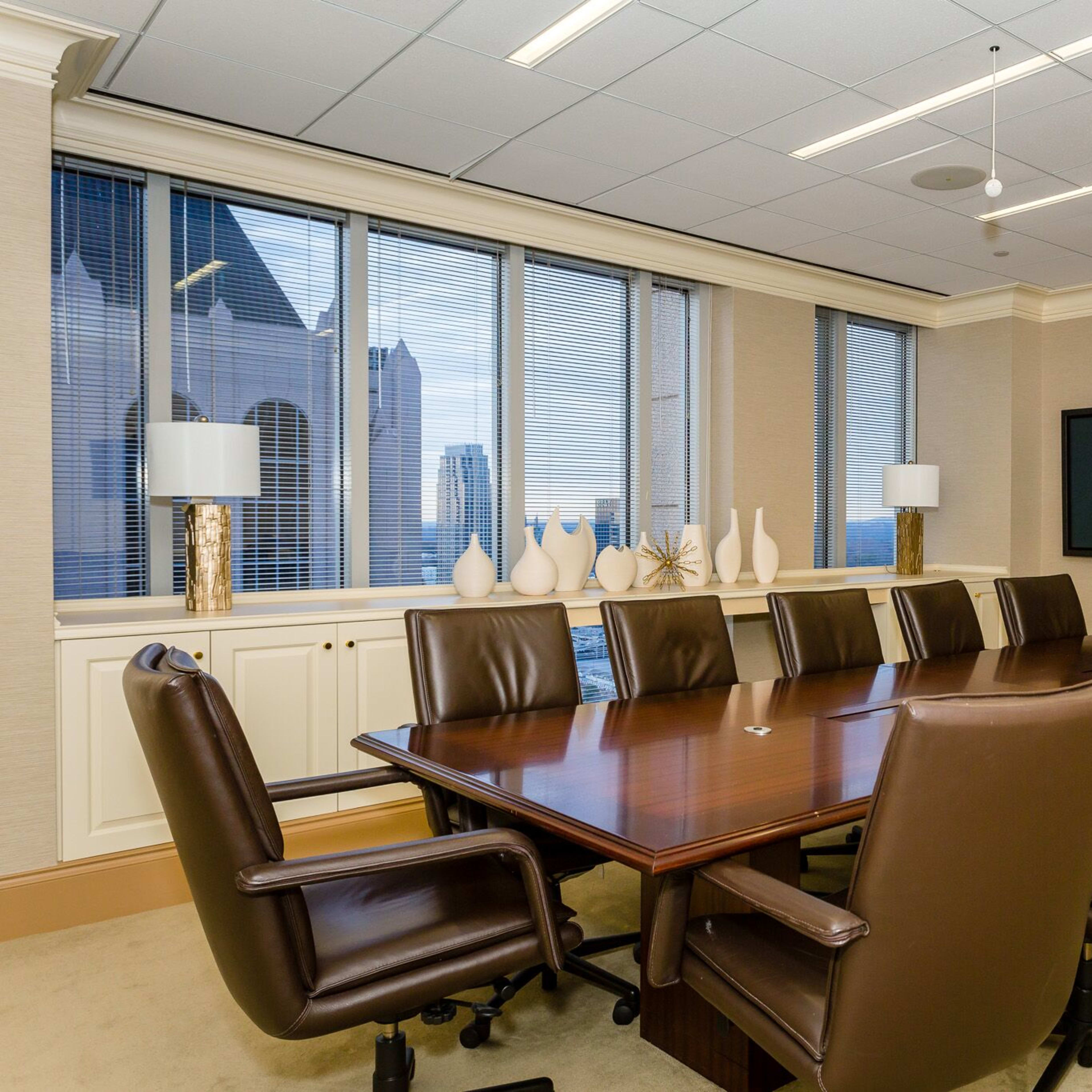 A spacious conference room features a large wooden table surrounded by leather chairs, with windows overlooking a cityscape and two wall-mounted screens.