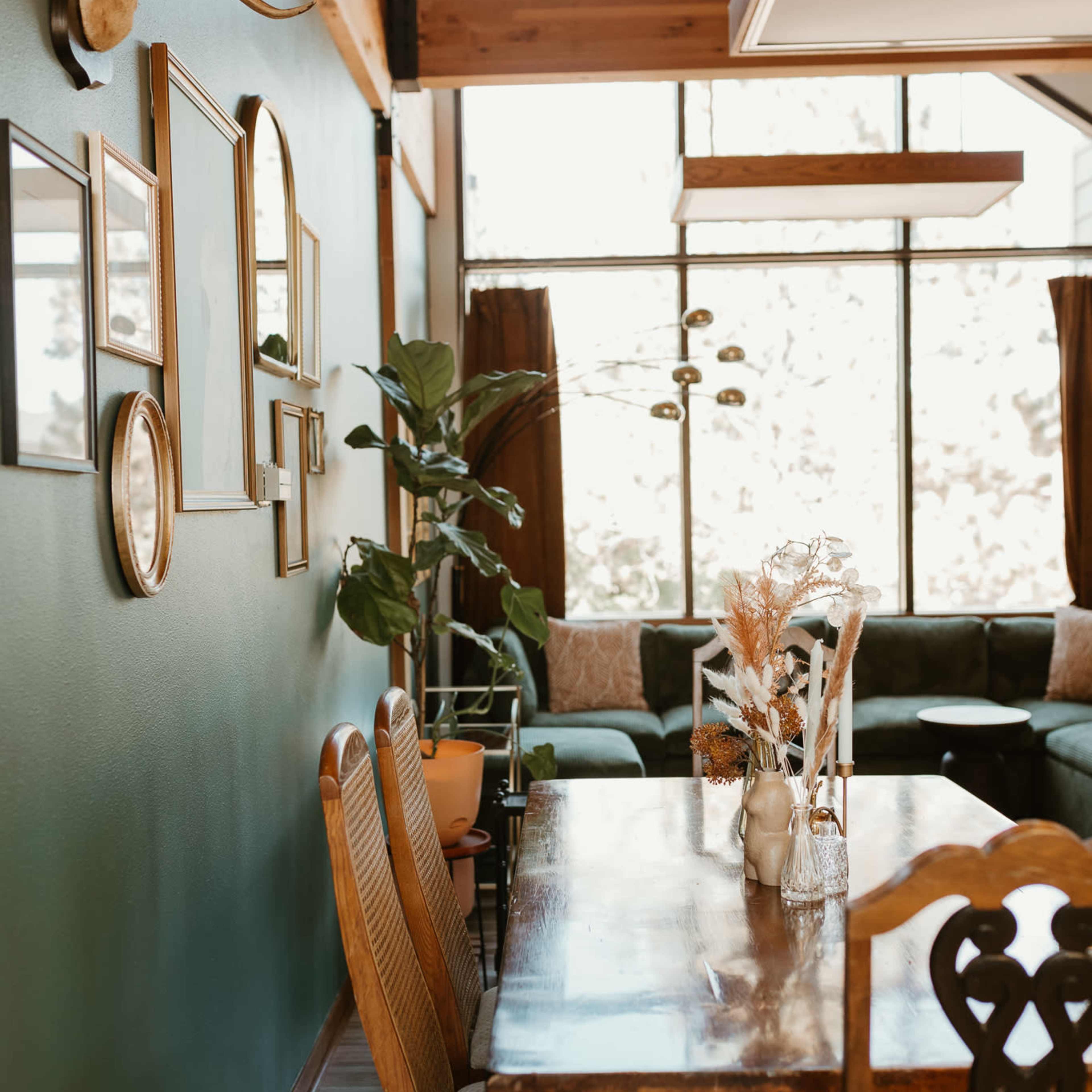 A dining area with a wooden table and chairs, side by side with a green wall featuring framed pictures and a decorative plant, leading to a spacious living area with large windows.