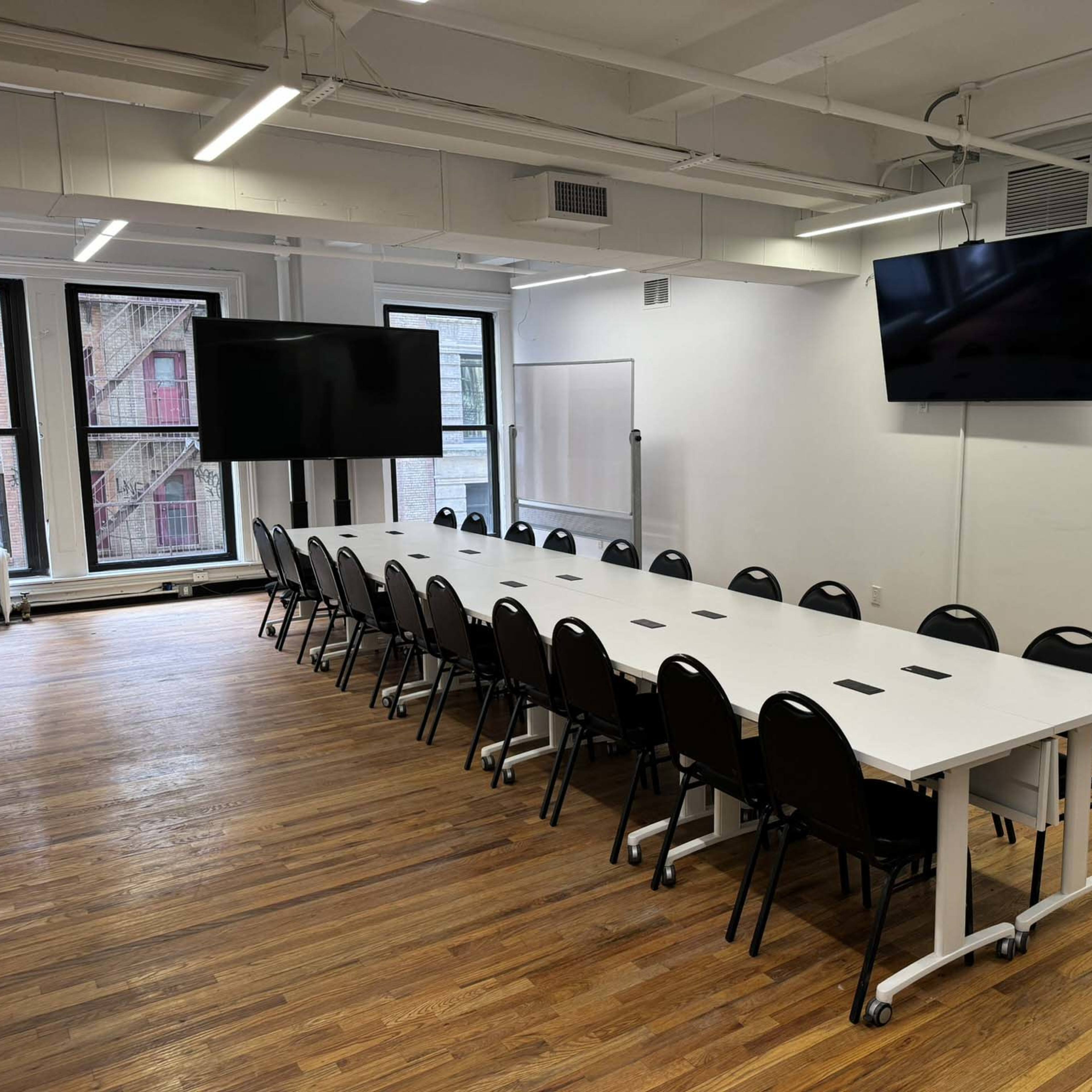 A long conference table with black chairs is arranged in a well-lit meeting room featuring two wall-mounted screens.