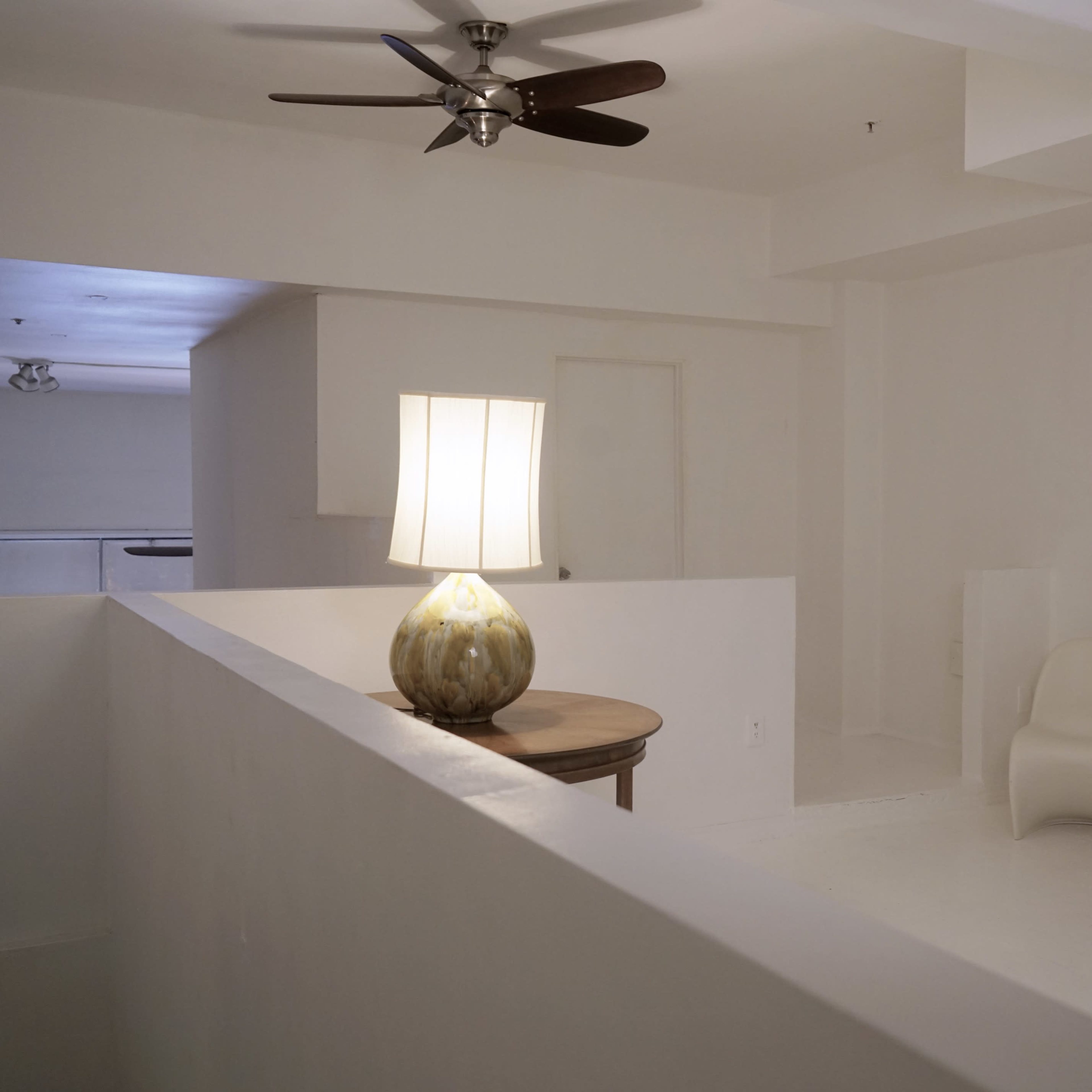 A minimalist interior features a lamp on a table, two white chairs, and a ceiling fan in a space with white walls and flooring.