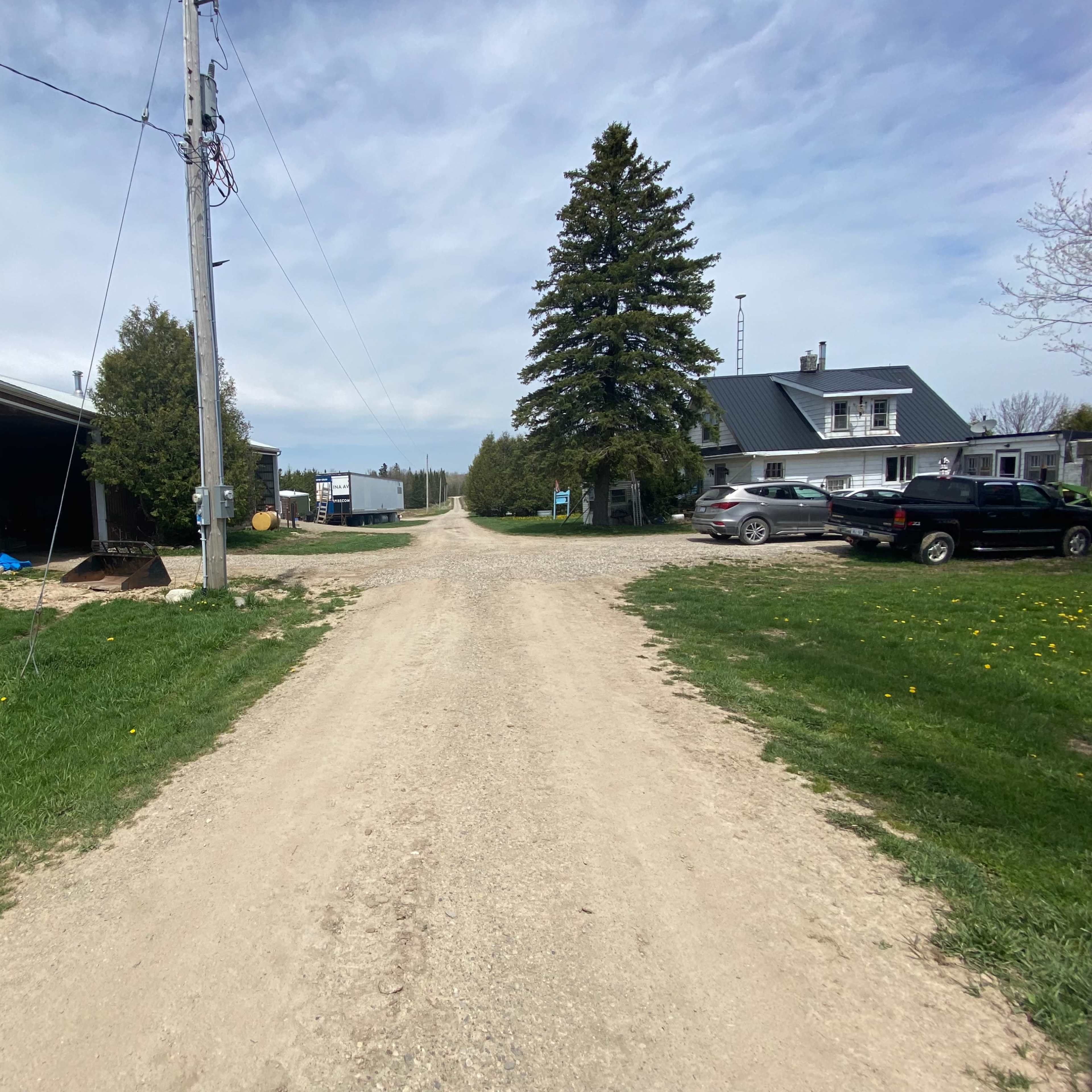 A dirt road leads through a rural area with two buildings on either side, flanked by trees and parked vehicles.