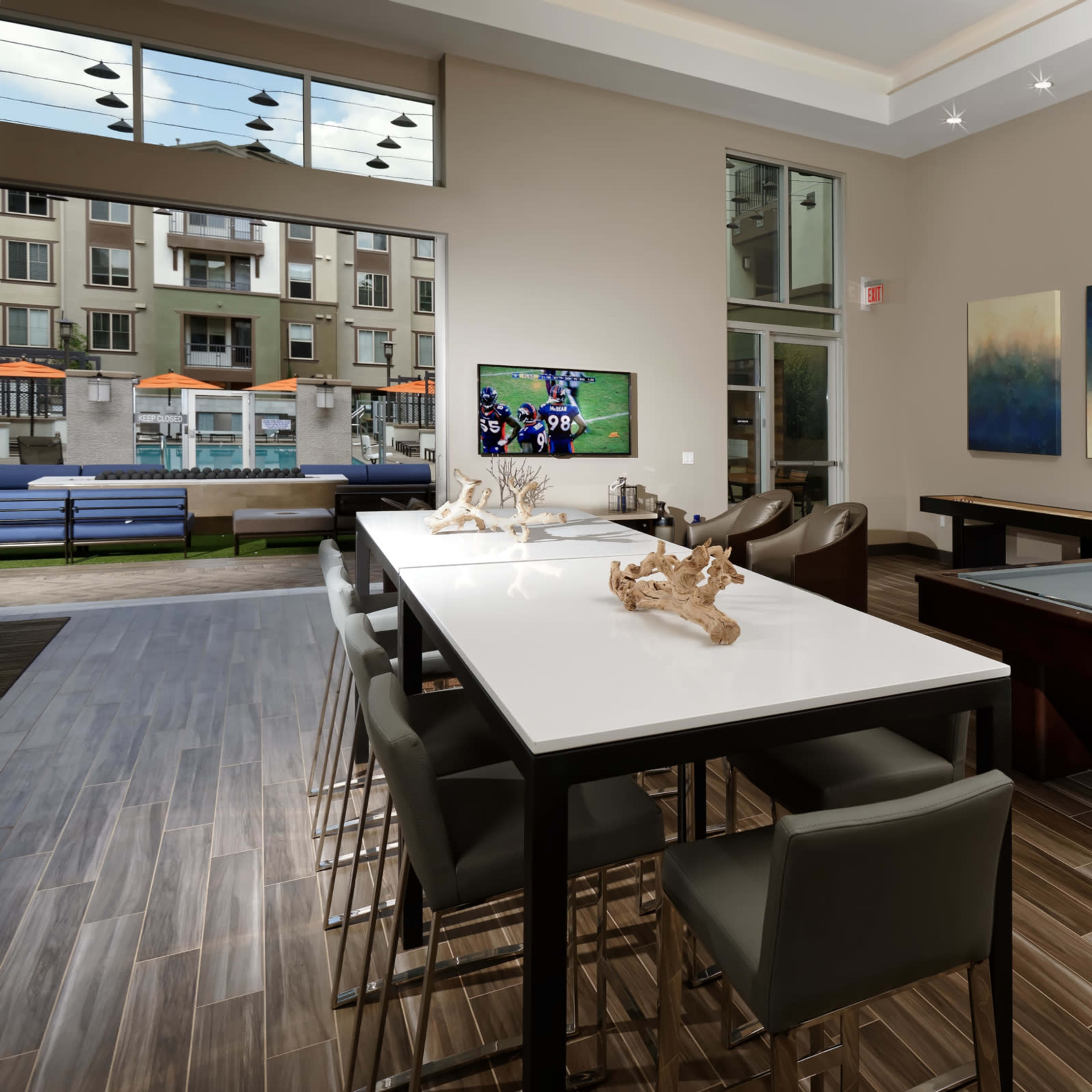 A modern lounge area featuring a pool table, a long dining table with chairs, and large windows overlooking an outdoor space with seating.