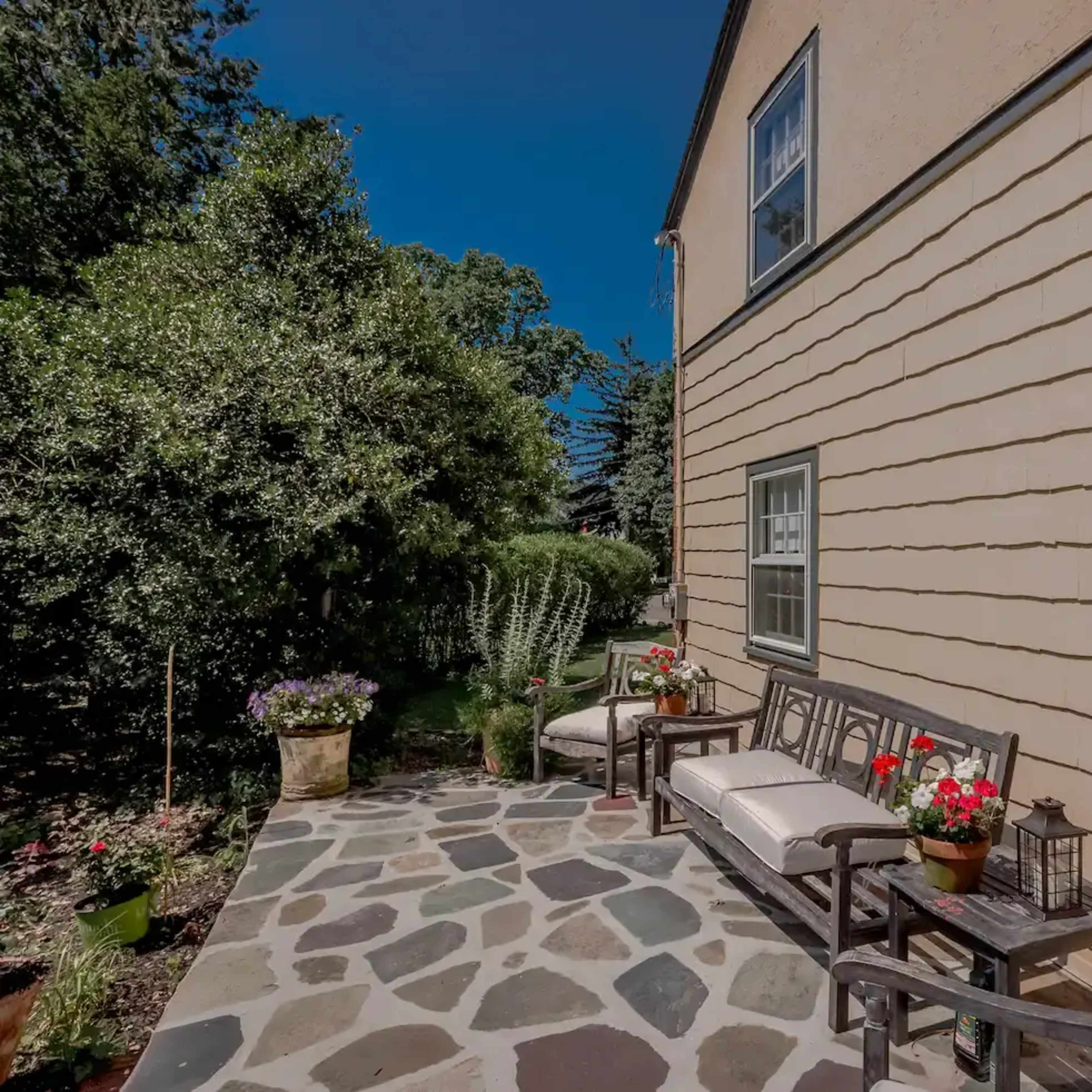 A stone patio with benches and flower pots is situated alongside a house, bordered by greenery.